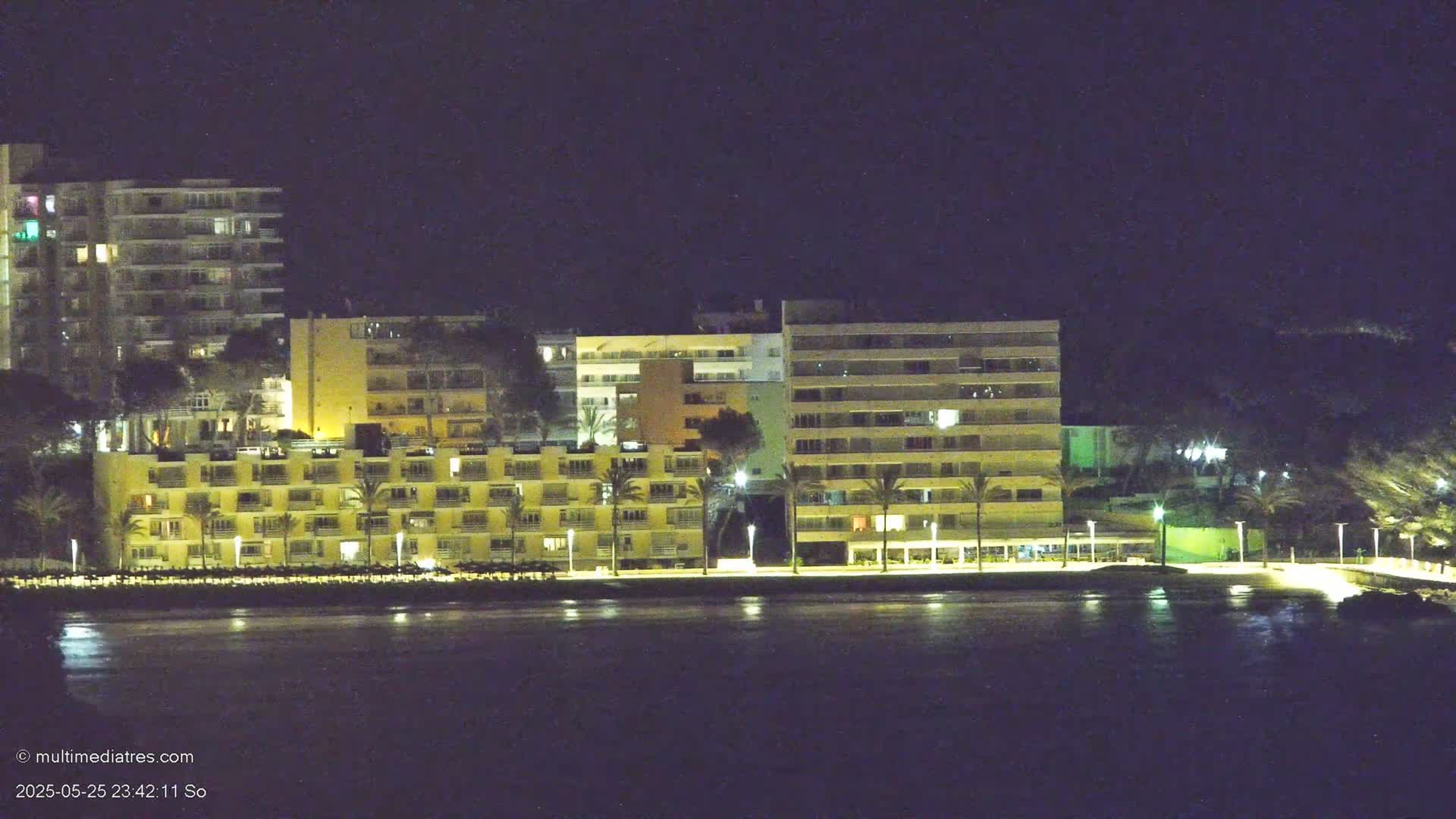 A row of illuminated apartment buildings sits along a calm body of water at night.