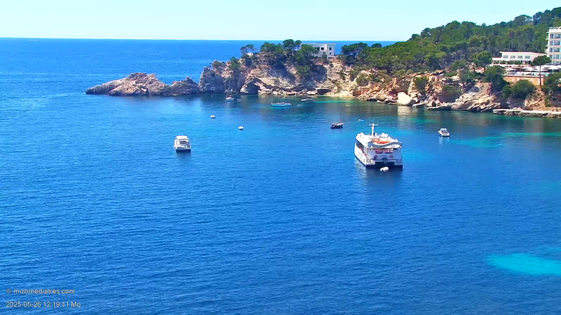 A calm, sunny day reveals a picturesque coastal scene with several boats in a clear blue bay, nestled amongst rocky cliffs and lush greenery.