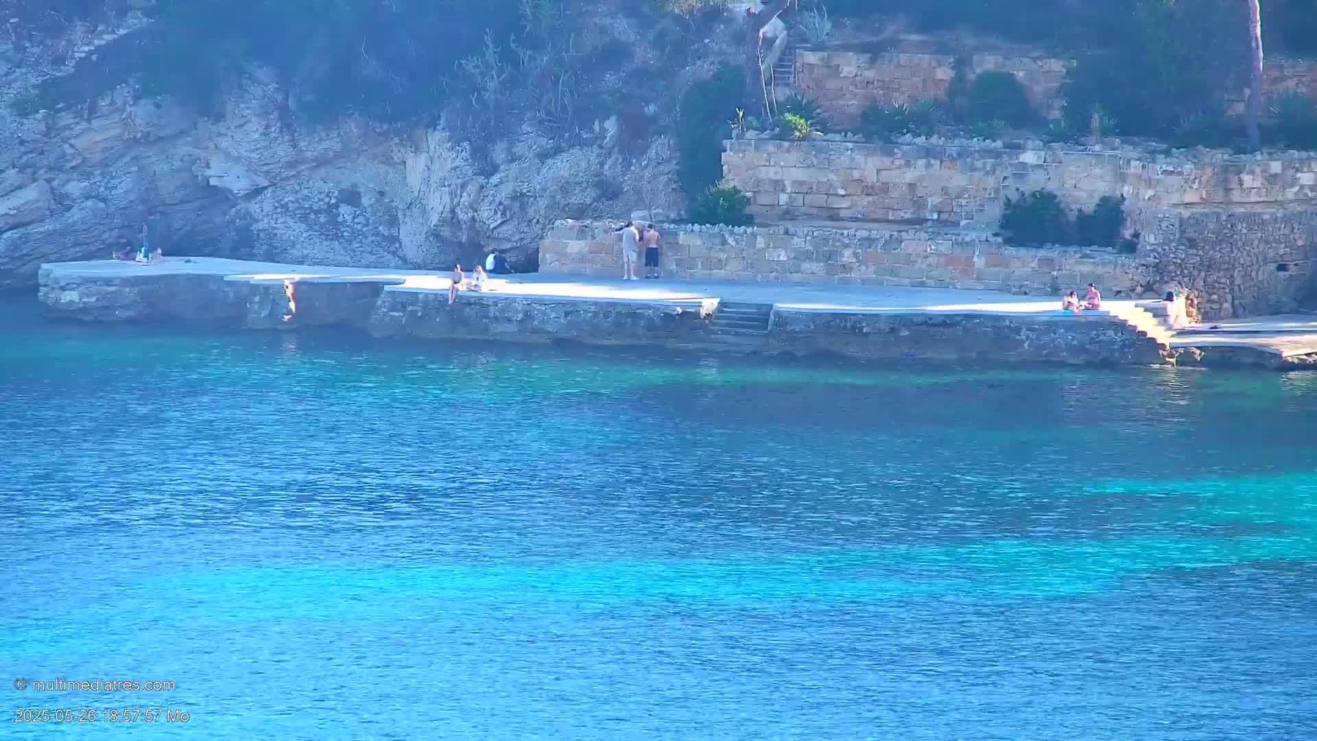 Several people relax on a stone platform overlooking a clear, blue body of water, next to a rocky cliffside.