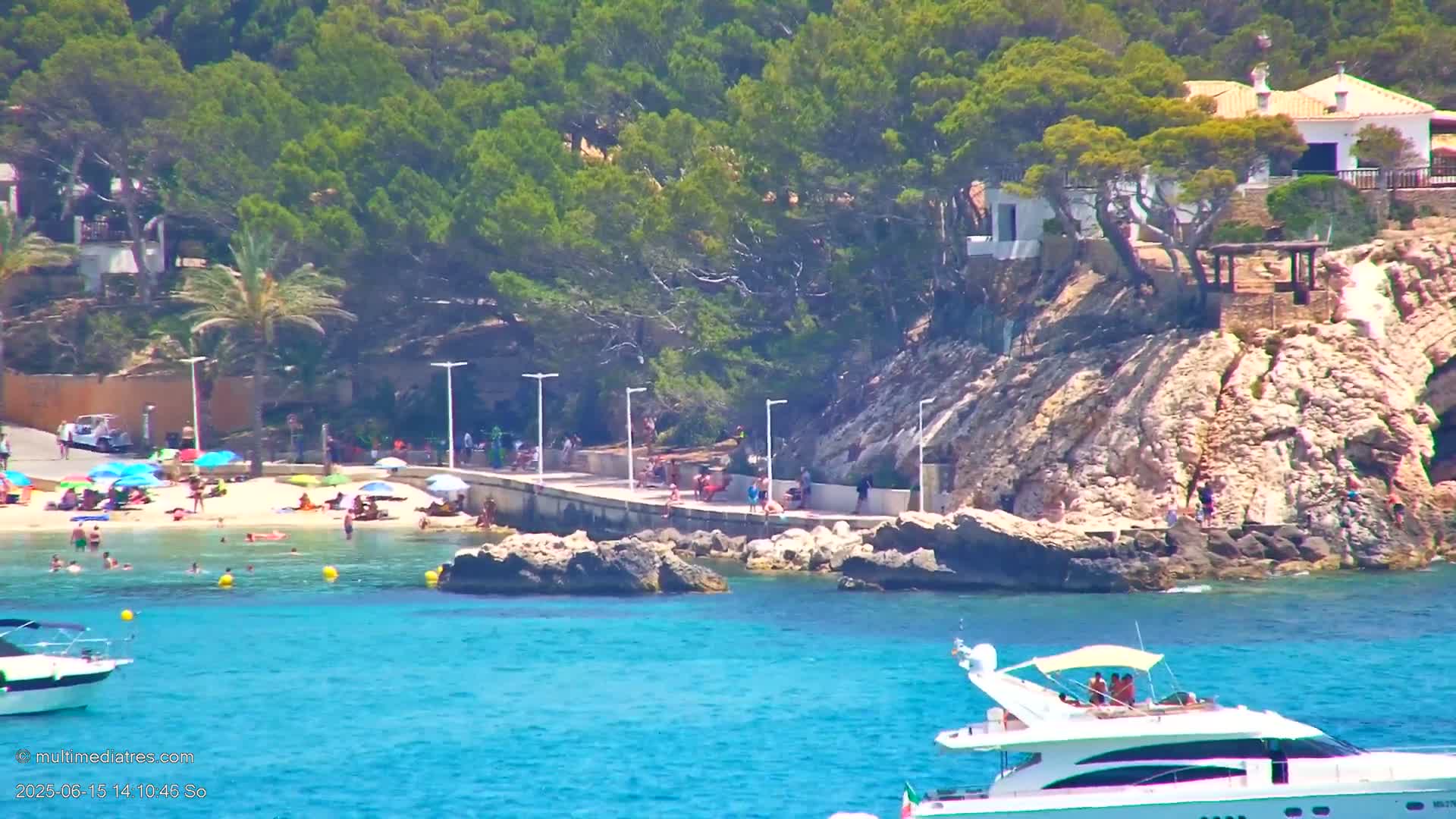 A sunny beach scene shows people relaxing on a sandy shore and swimming in clear turquoise water, with boats anchored offshore and houses perched on a rocky cliff overlooking the cove.
