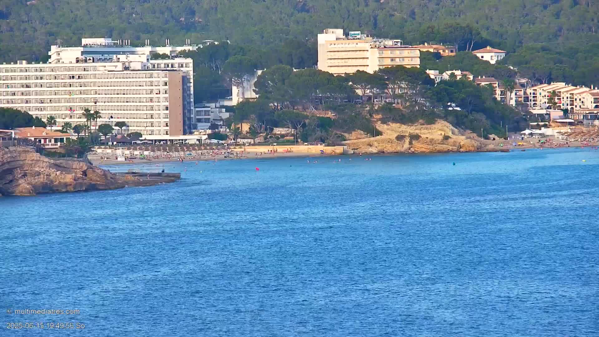 A calm, blue ocean stretches out in front of a beach with people sunbathing, backed by several large hotels and buildings nestled among green hills.