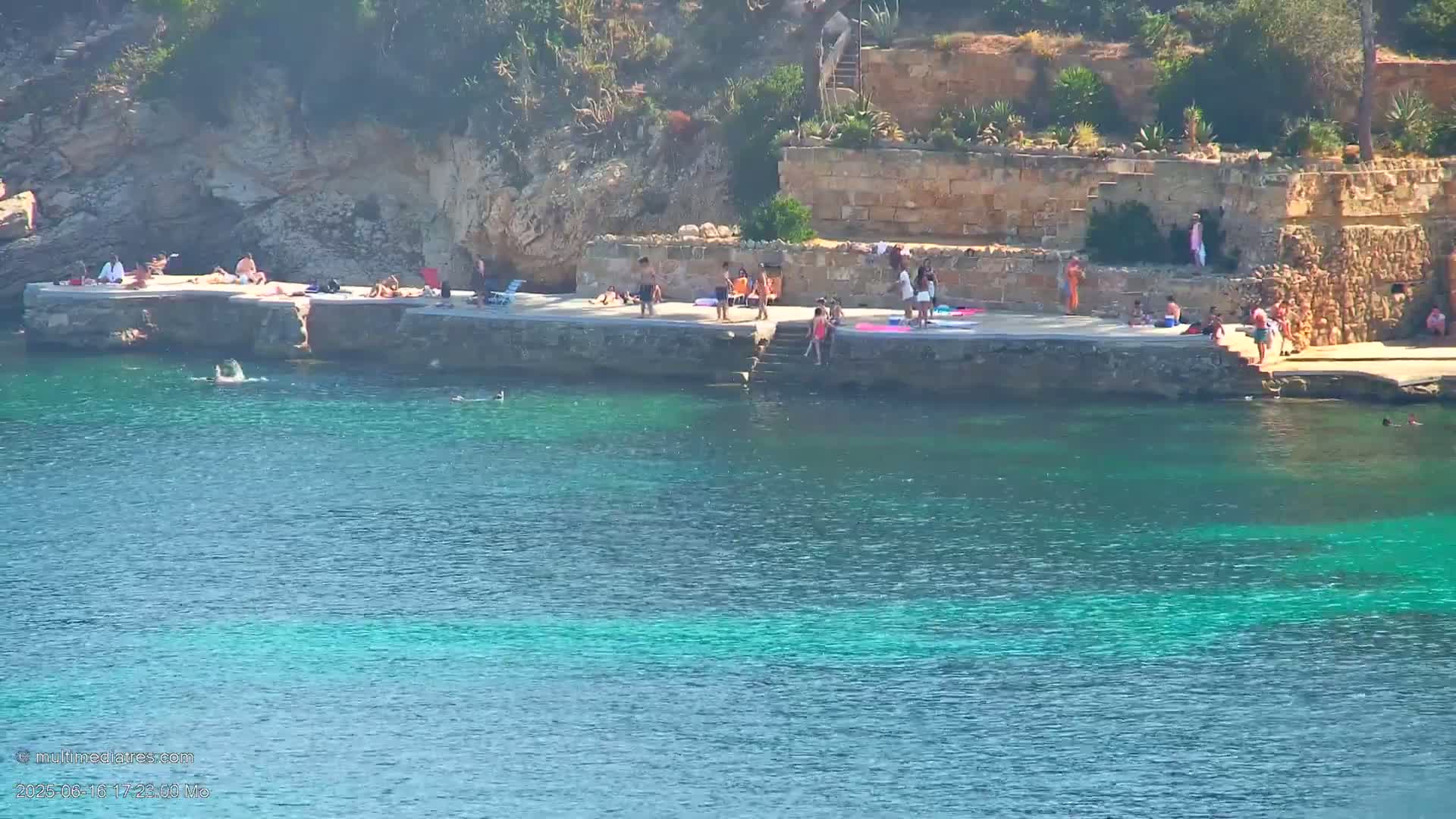 People are swimming and sunbathing on a rocky shore next to clear, turquoise water on a sunny day.