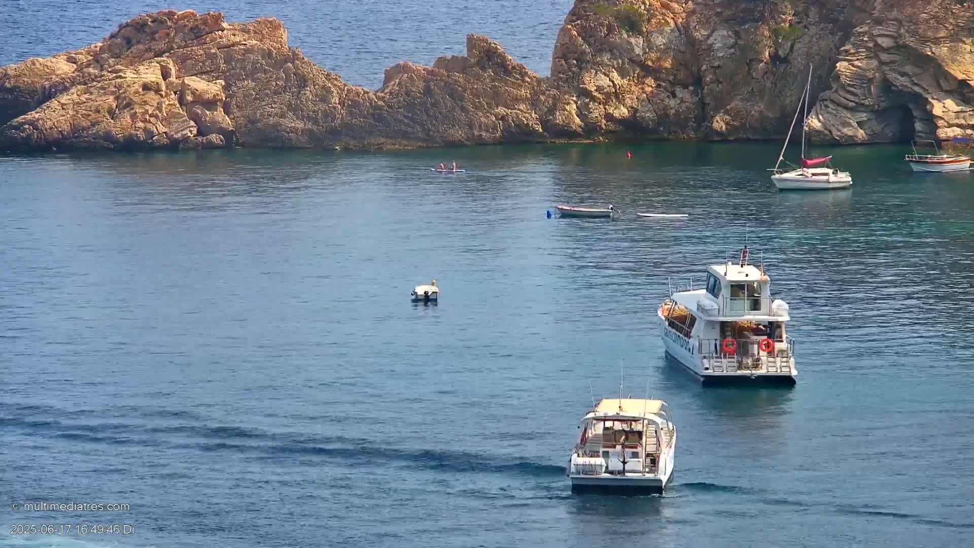 Calm blue water surrounds rocky outcrops and several small boats, including two passenger vessels.