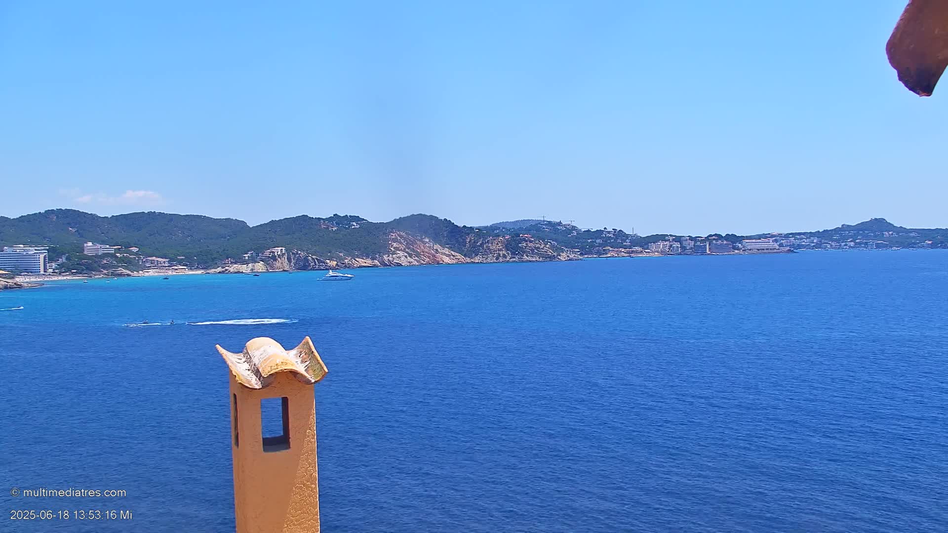 A clear blue ocean stretches to a distant coastline of green hills and low buildings under a bright blue sky, partially visible behind a tan chimney.