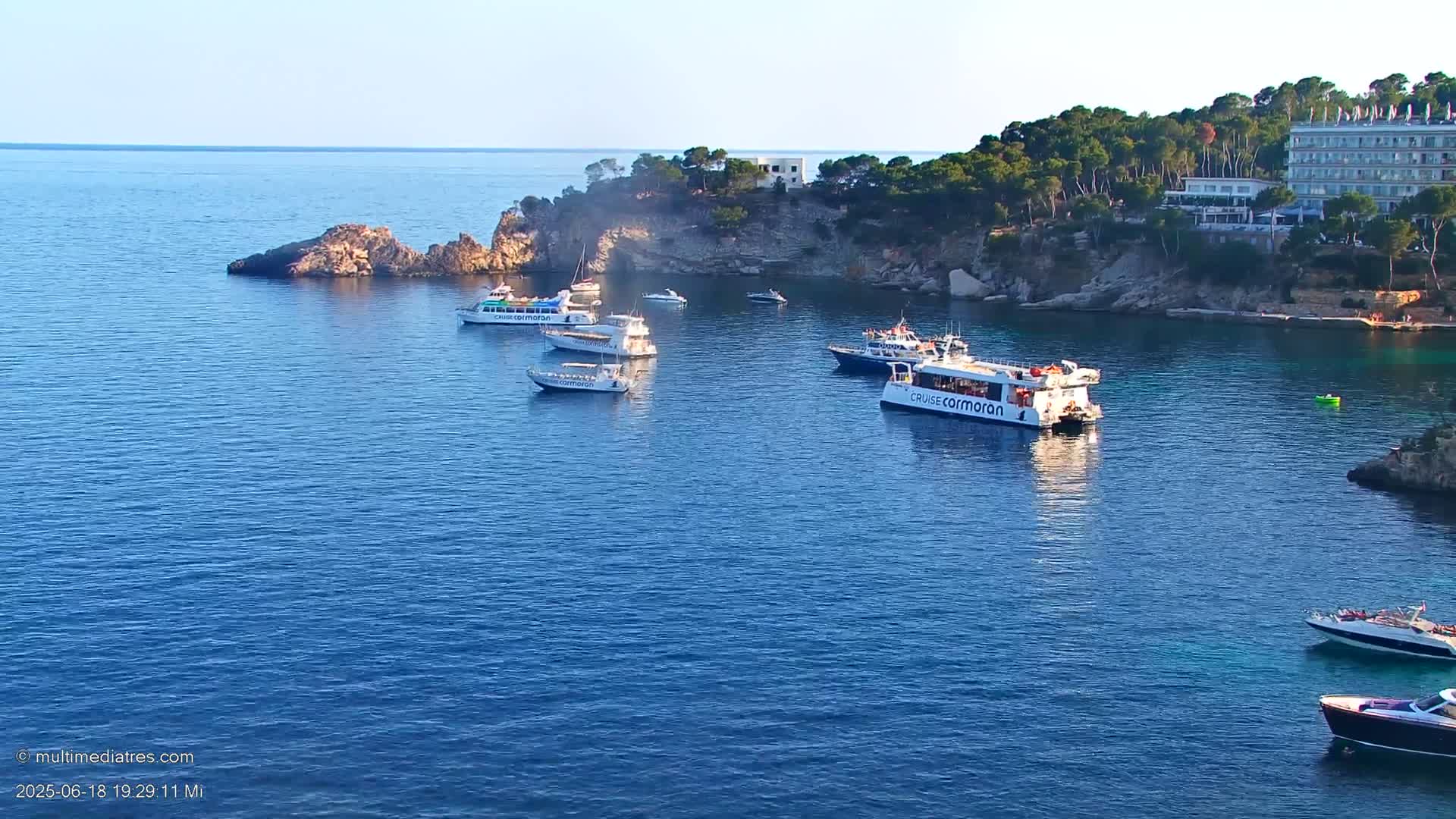 Several boats are moored in a calm, blue bay next to a rocky coastline with green vegetation and a building on top.