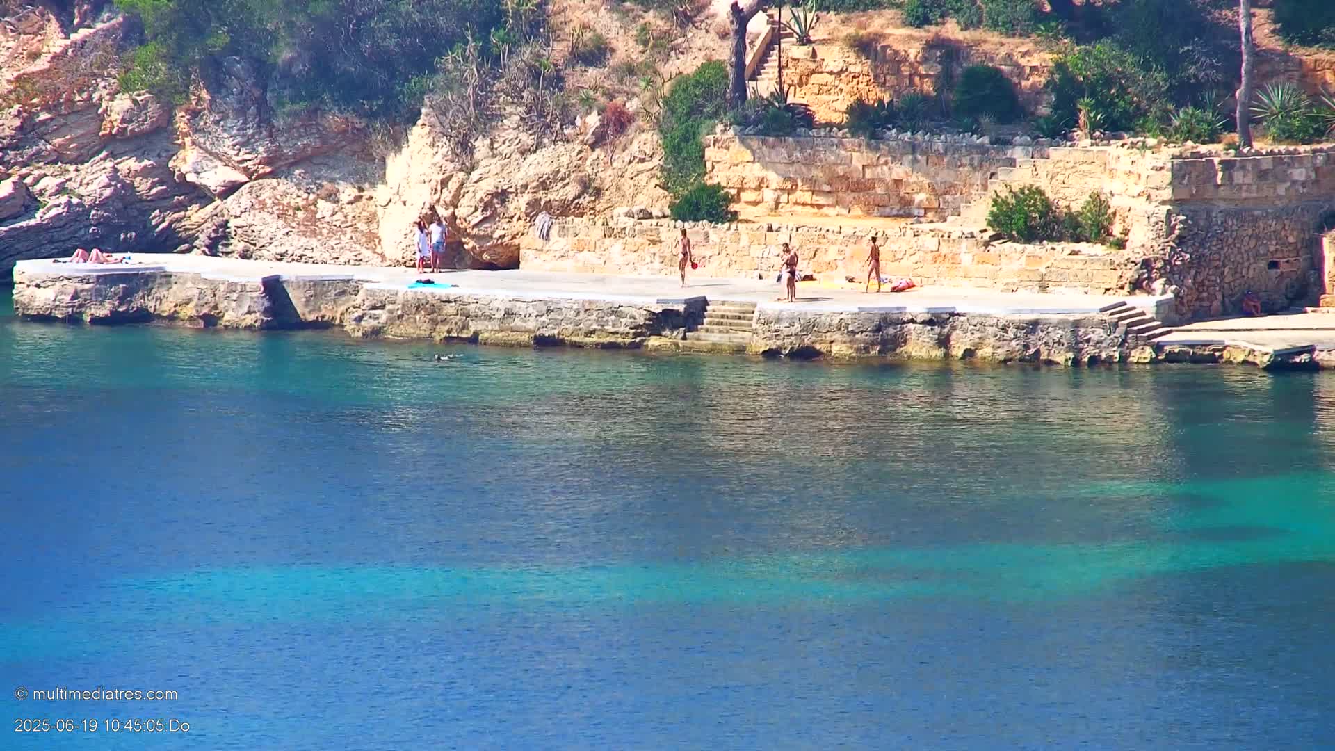 Several people are sunbathing and swimming at a rocky, seaside location with clear, blue-green water on a sunny day.