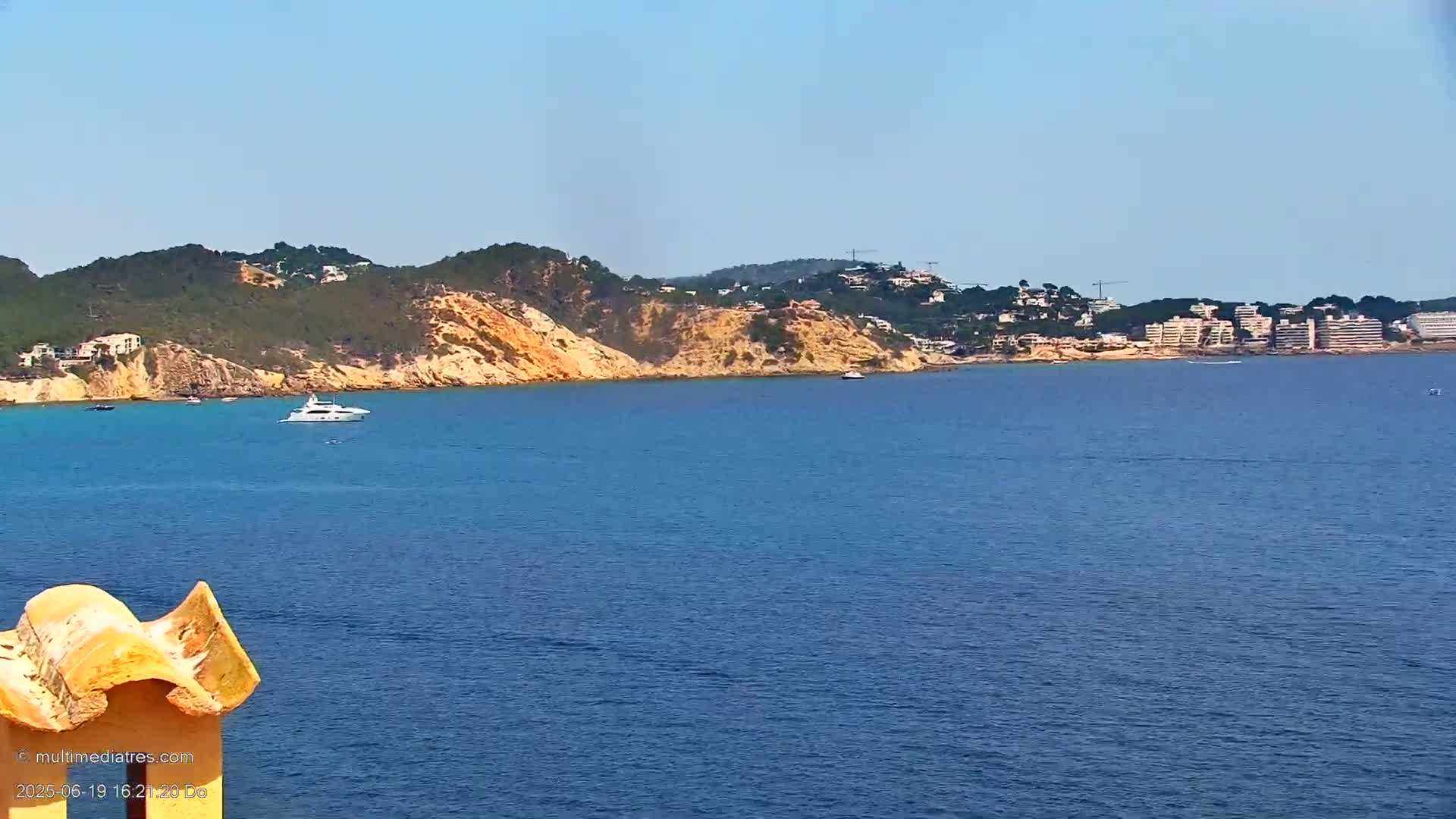 A yacht is visible on a calm, blue sea, with a hilly coastline featuring buildings and vegetation in the background under a clear sky.