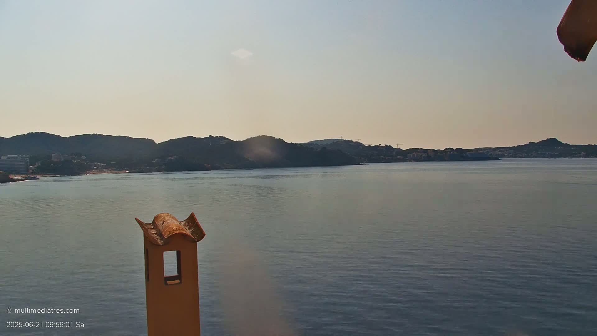 A calm body of water stretches out to a distant shoreline of hills and buildings under a clear sky.