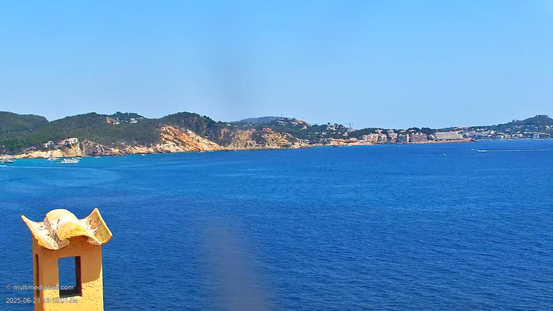 A view from a chimney shows a calm blue ocean with a distant coastline of green hills, buildings, and a few boats on a sunny day.