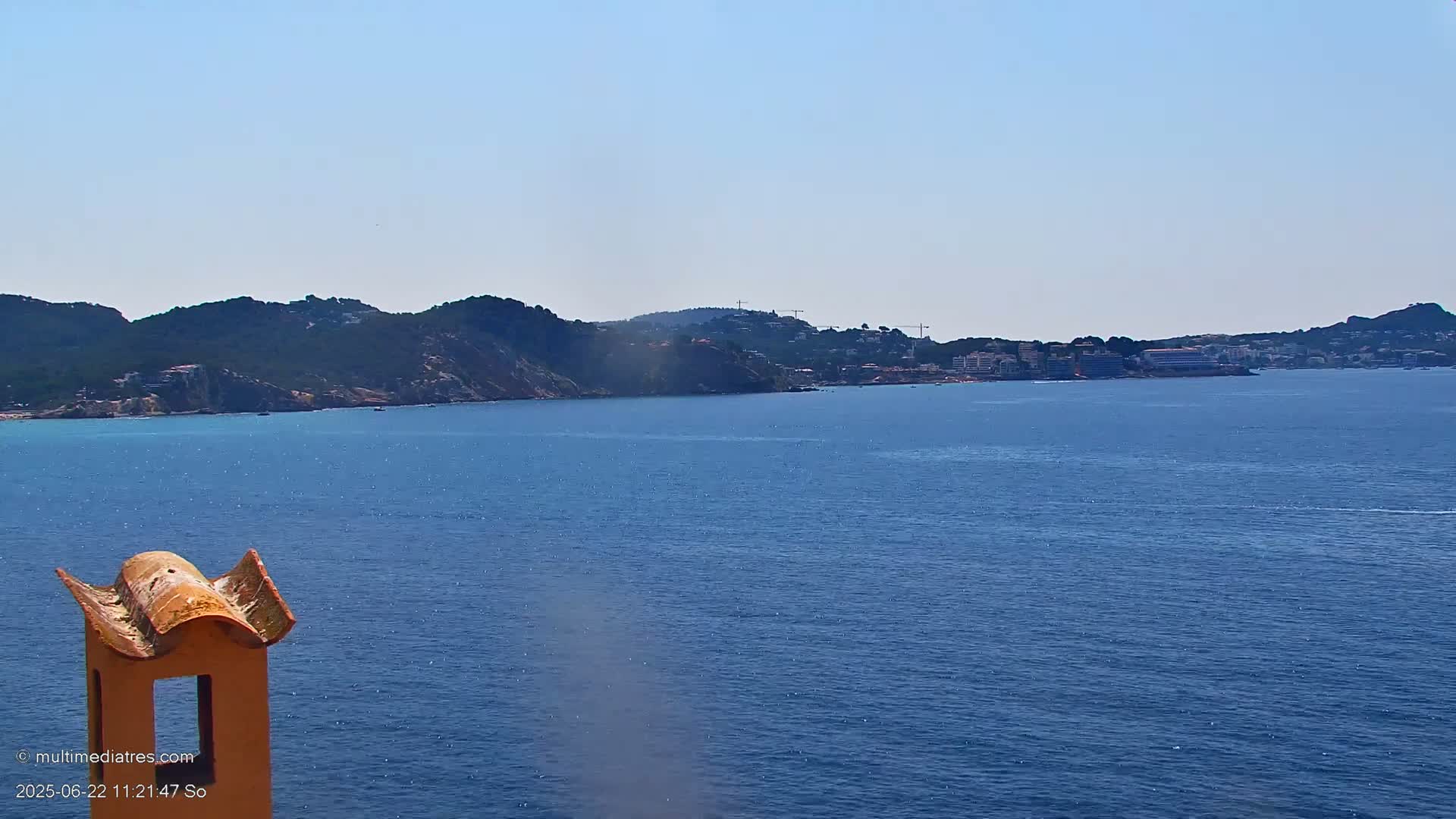 A view from a rooftop shows a calm, blue ocean separating a rocky, green hillside from a distant coastline with buildings under a clear sky.