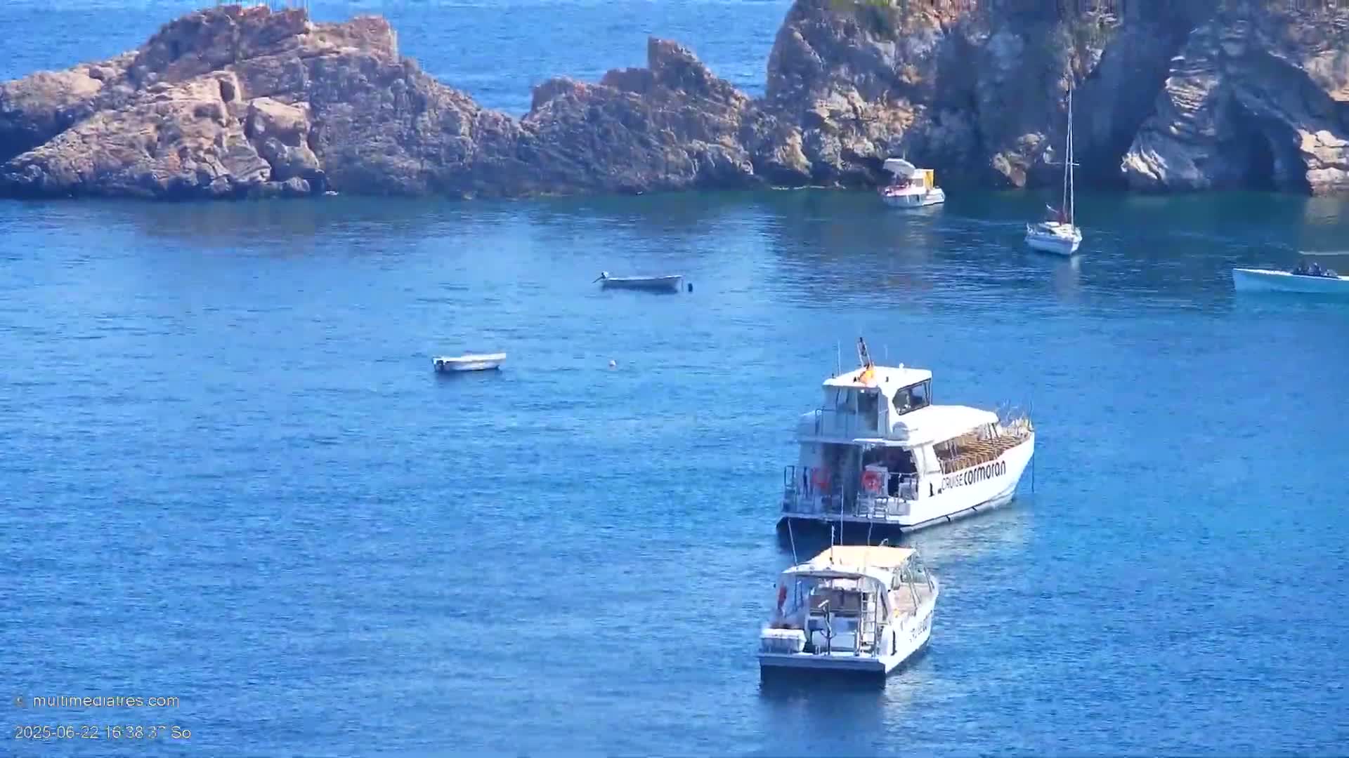 Several boats are anchored in calm, clear blue water near rocky islands on a sunny day.