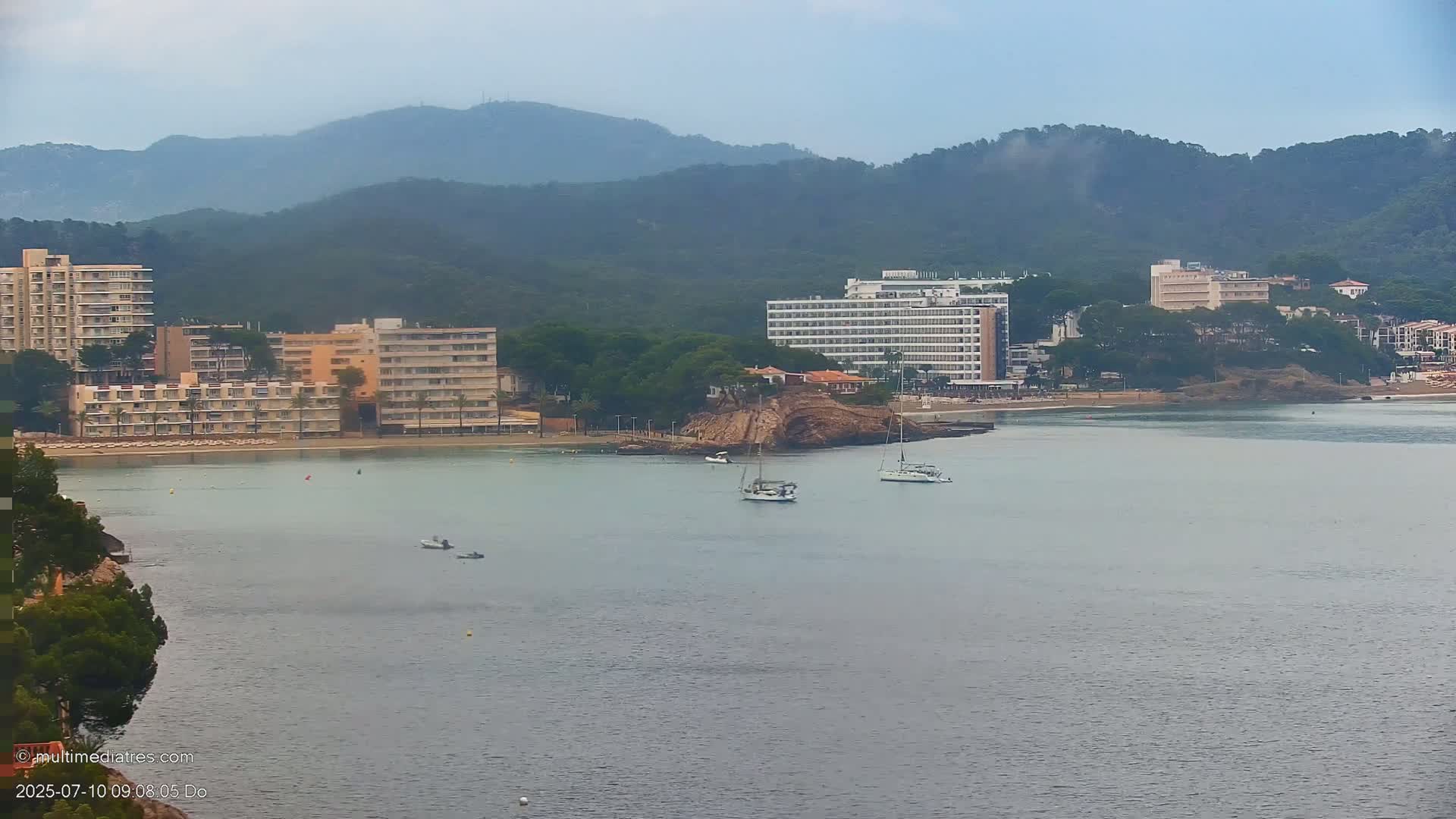 Several hotels line a calm bay under a hazy sky, with a few boats anchored in the water and hills in the background.
