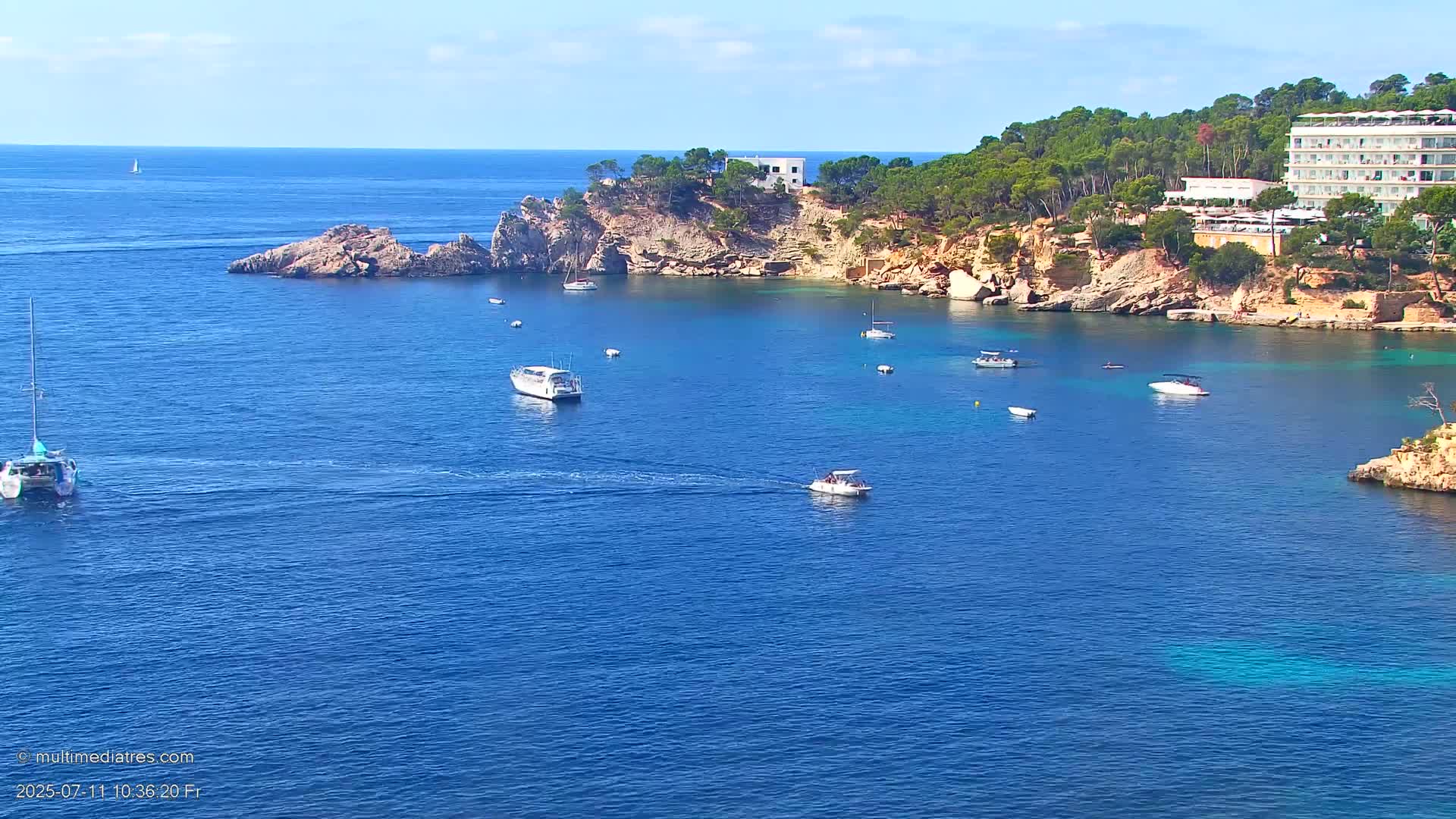 A calm, sunny day reveals a secluded cove with numerous boats, a rocky shoreline lined with lush greenery and a large building, all surrounded by deep blue water.