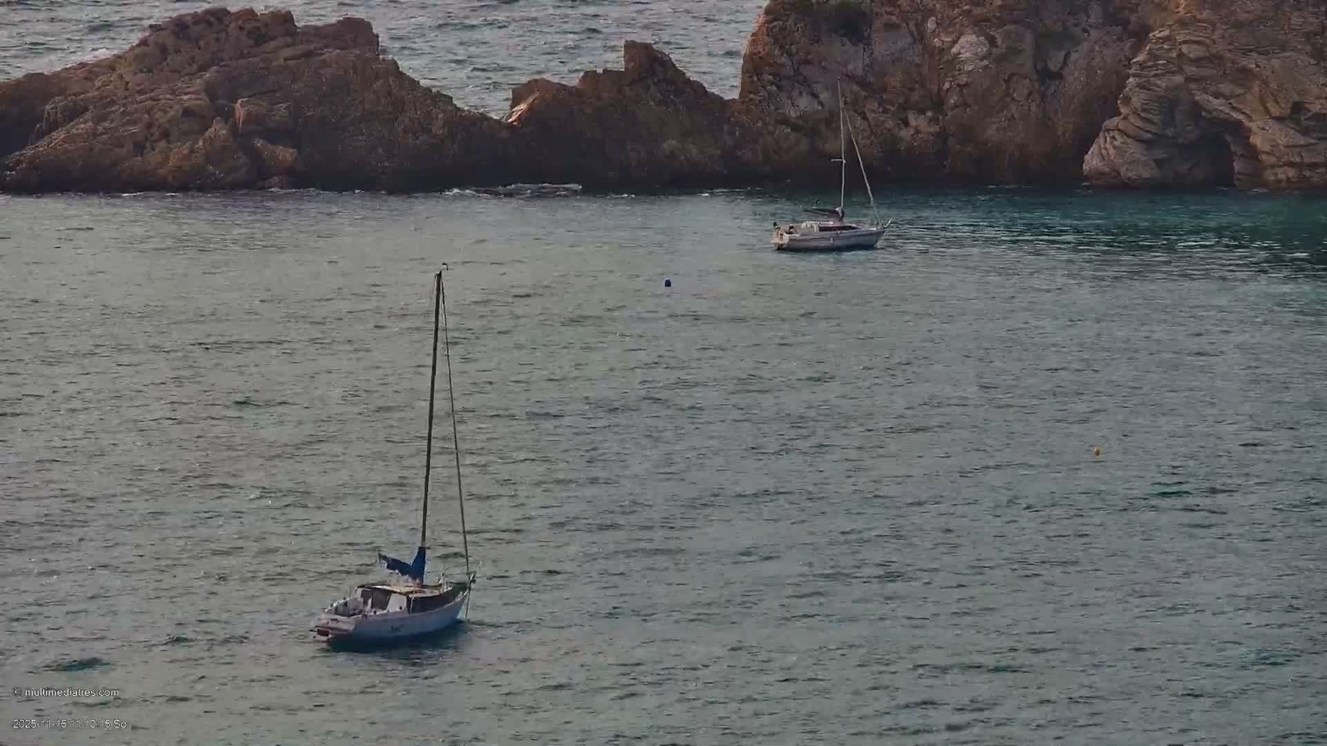 Two sailboats gently float on calm, rippled waters near a rugged rocky coastline under clear daytime conditions.
