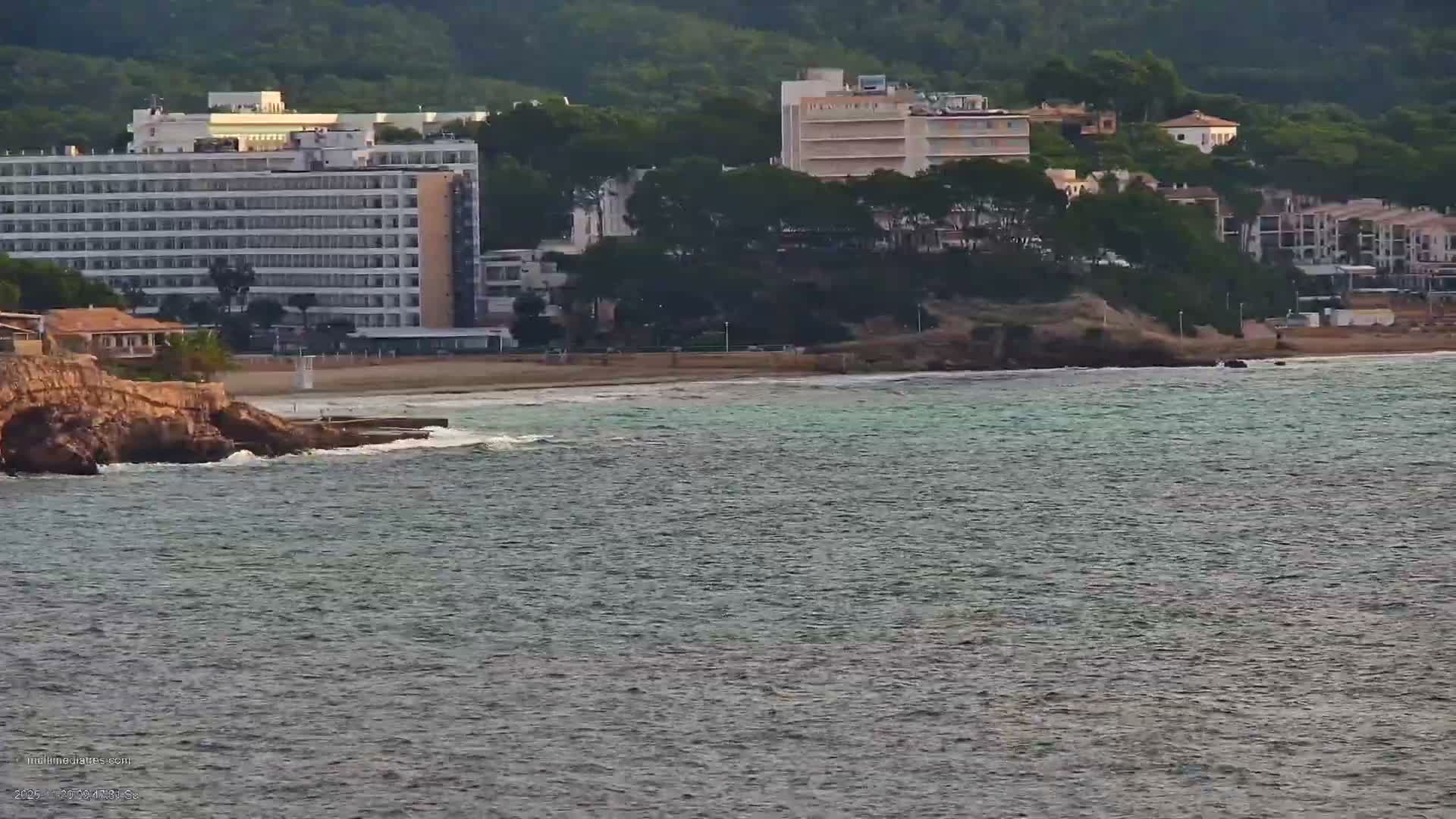 Under an overcast sky, a tranquil grey-blue sea with gentle waves washes upon a rocky shore and sandy beach, backed by numerous large buildings and lush green hills.