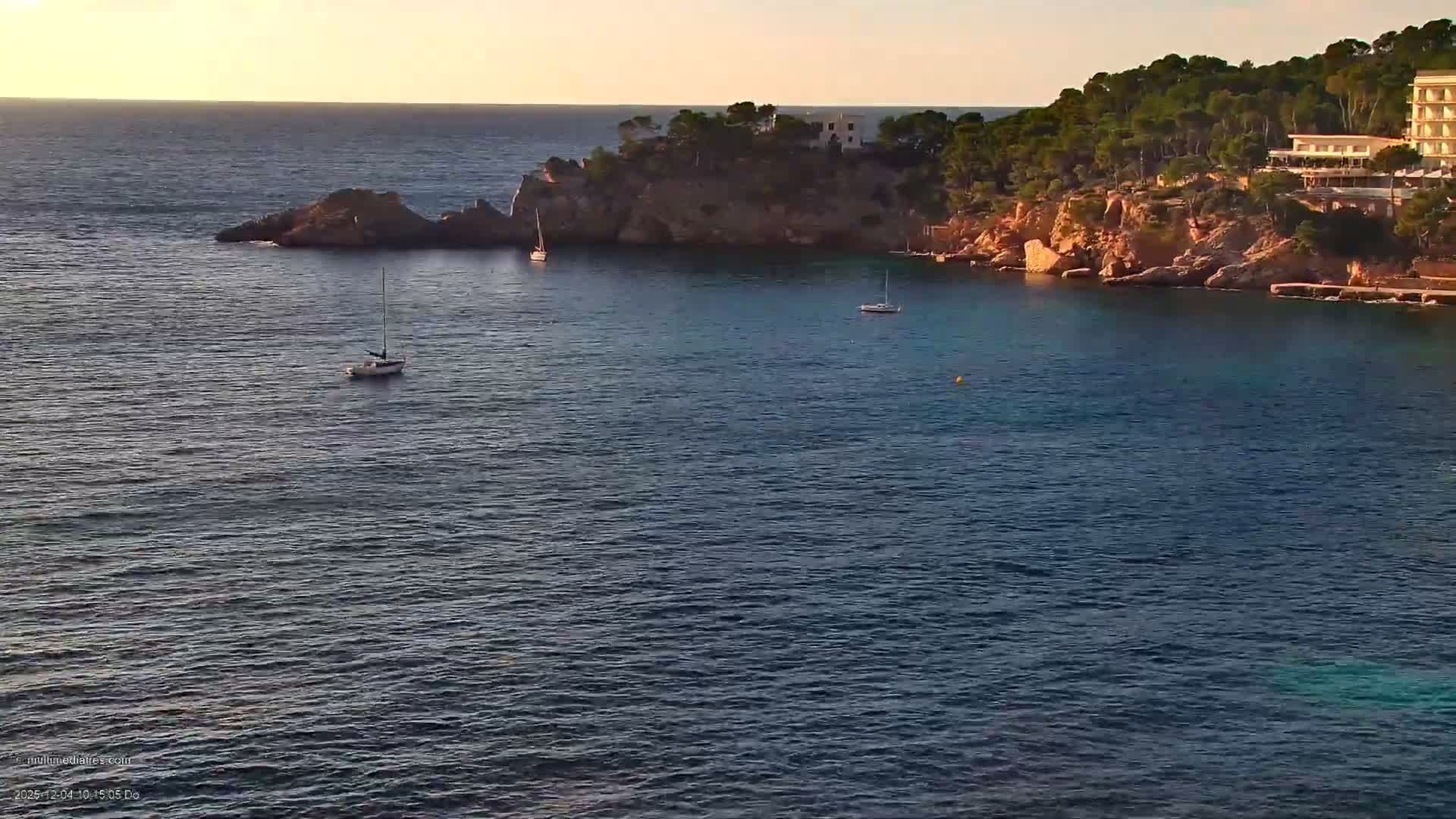 The image shows a tranquil ocean bay with two sailboats, backed by a lush, tree-covered rocky coastline featuring buildings, all under a clear and sunny sky.