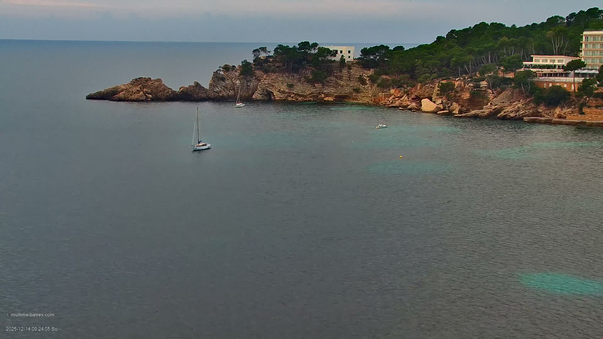 The image shows a tranquil ocean bay with two sailboats, backed by a lush, tree-covered rocky coastline featuring buildings, all under a clear and sunny sky.