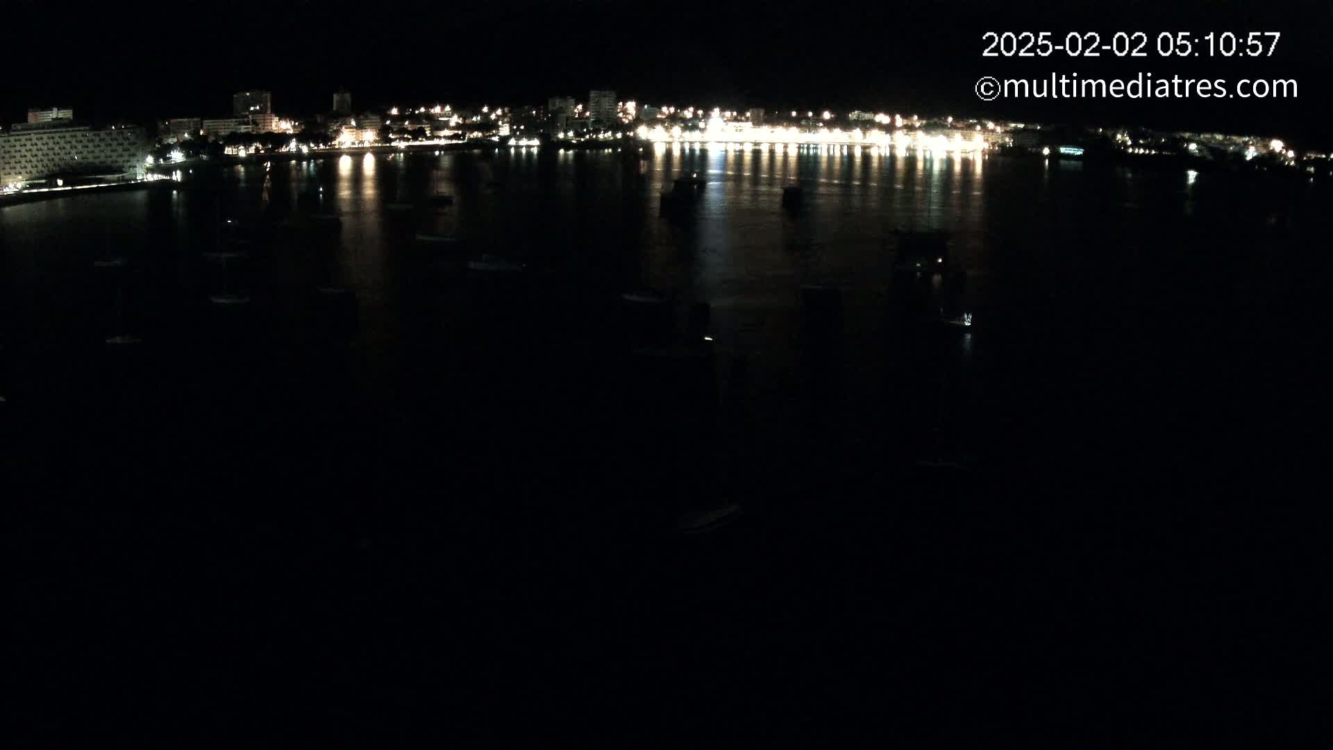 A nighttime view of a city's illuminated waterfront, showing numerous boats moored in dark water.