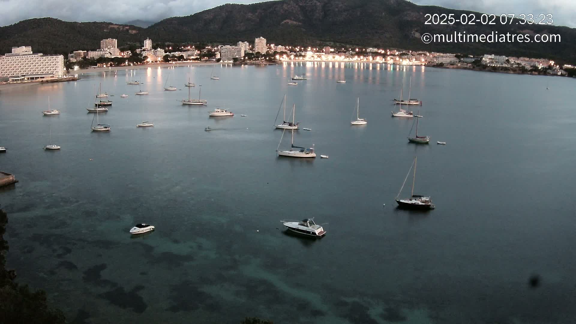Many sailboats are anchored in a calm bay at twilight, with a town and hotels illuminated along the shore backed by dark mountains.