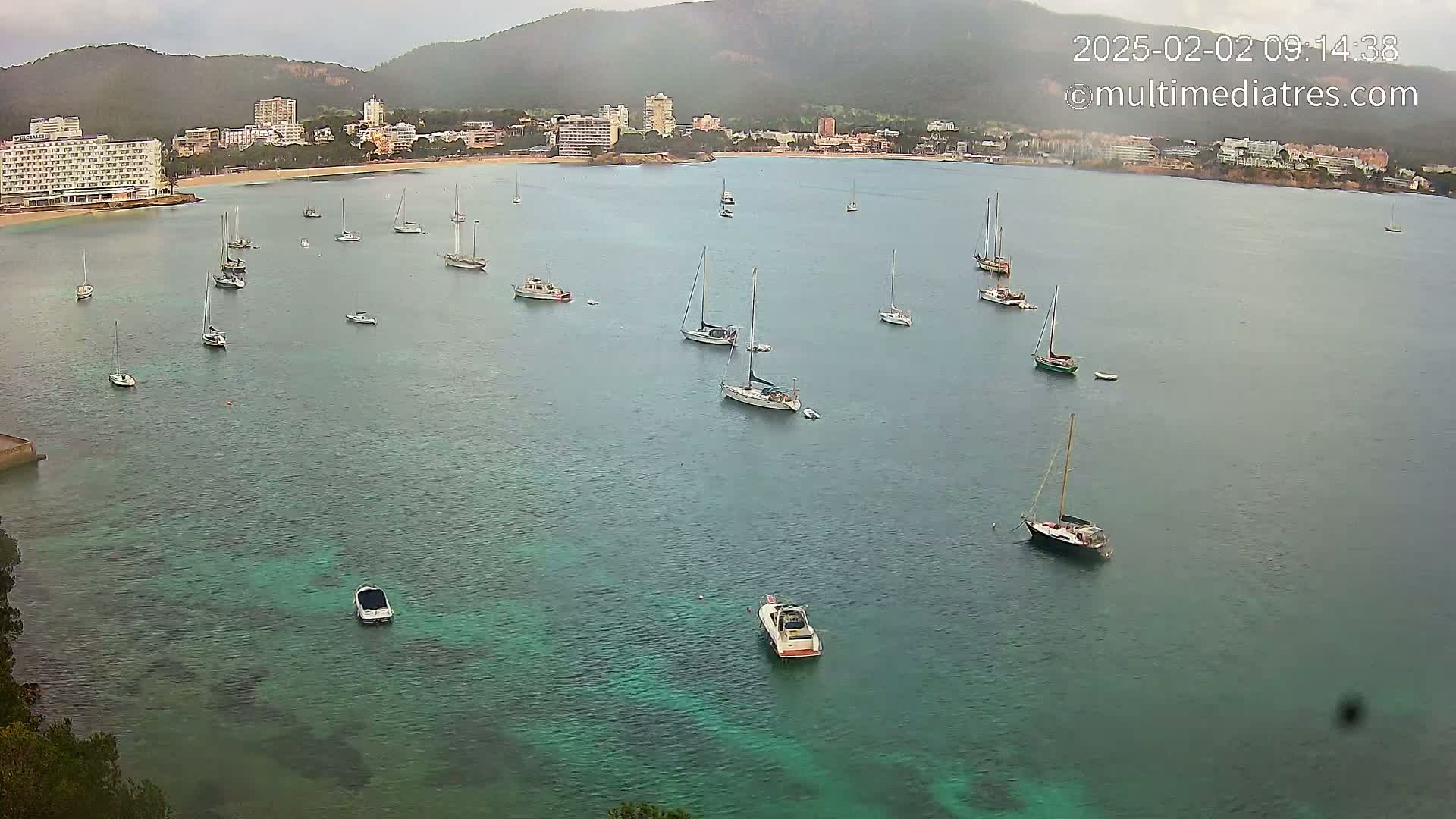 A calm, overcast day reveals numerous sailboats and motorboats anchored in a bay near a shoreline with buildings and hills in the background.