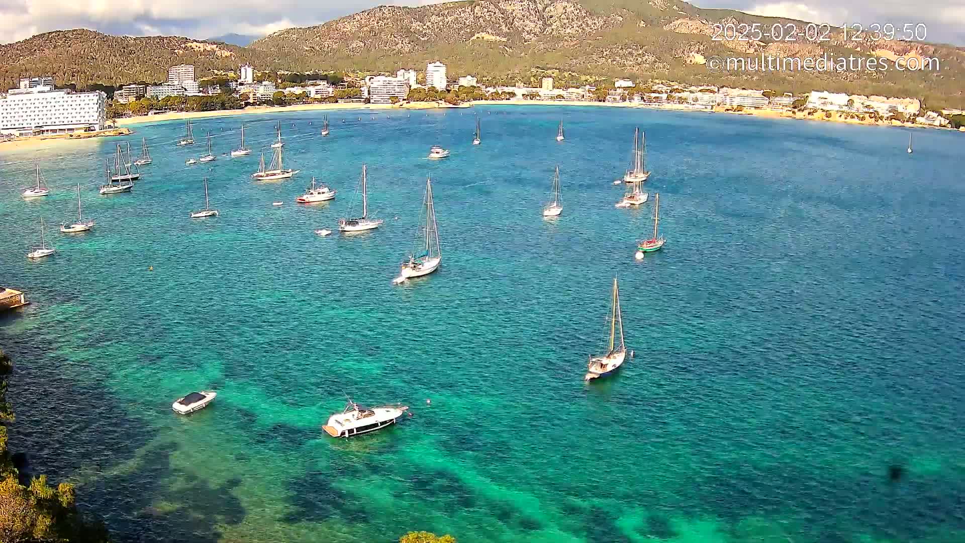 Numerous sailboats and motorboats are anchored in a calm, clear, turquoise bay, with a mountainous coastline and buildings in the background under partly cloudy skies.