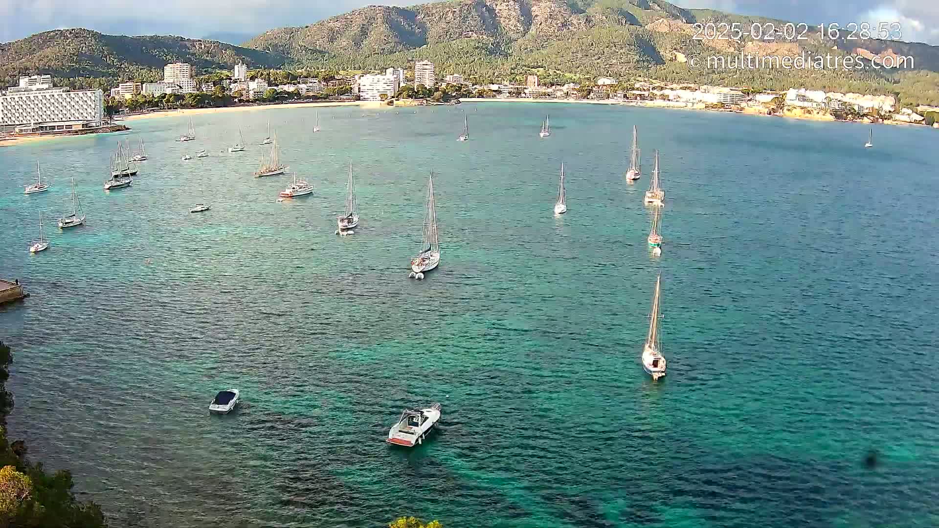 A calm, clear turquoise bay with numerous sailboats and a few motorboats anchored near a beach and town nestled against a mountain range under a mostly sunny sky.
