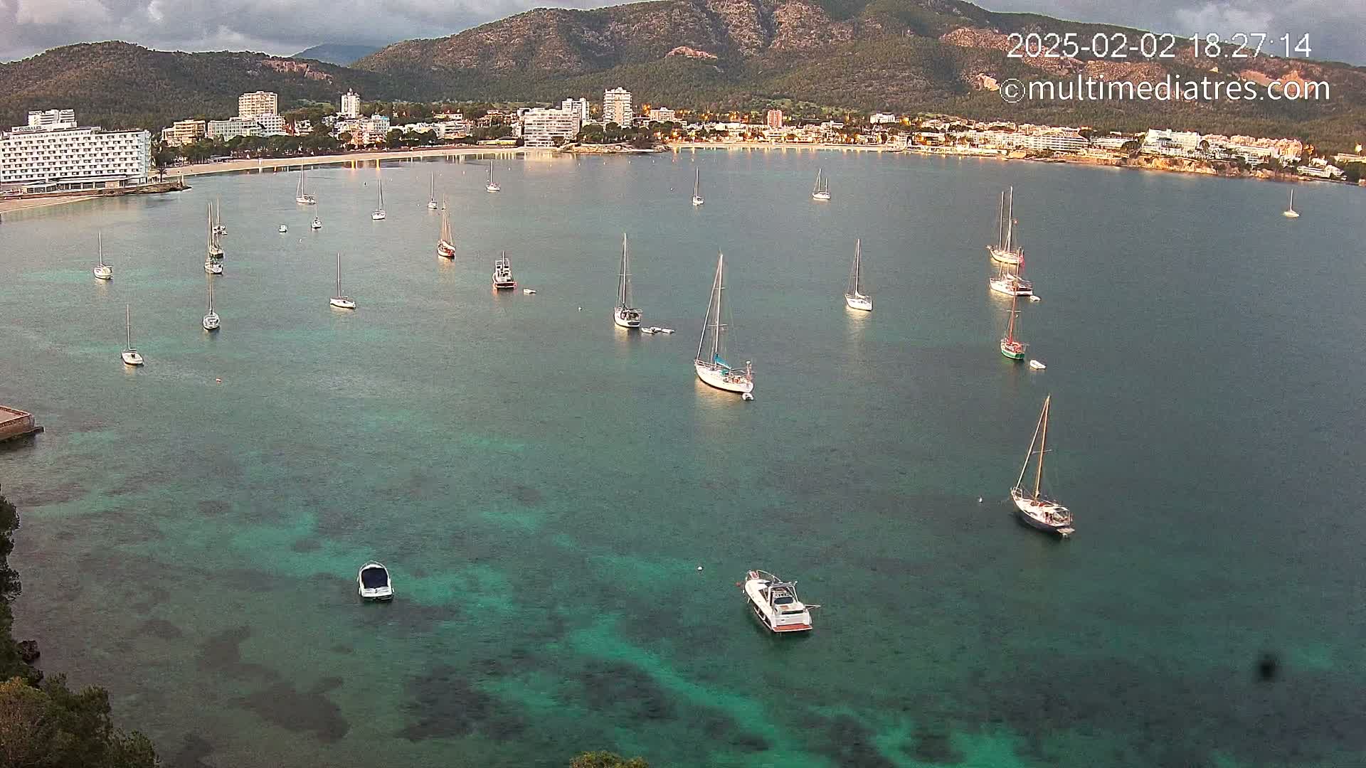 Numerous sailboats are anchored in a calm, teal bay near a coastline with hotels and mountains under a partly cloudy sky.
