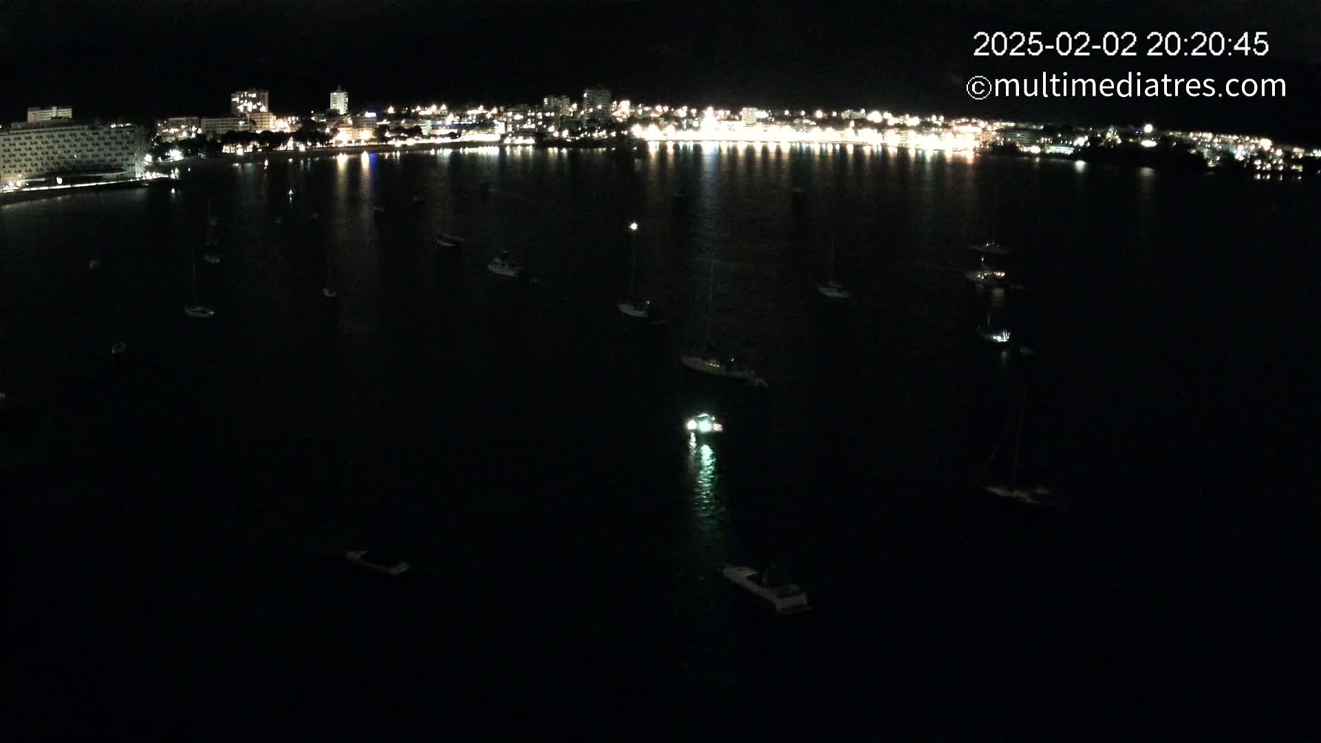 A nighttime, dark-water harbor scene shows many small boats and a brightly lit city skyline in the distance.