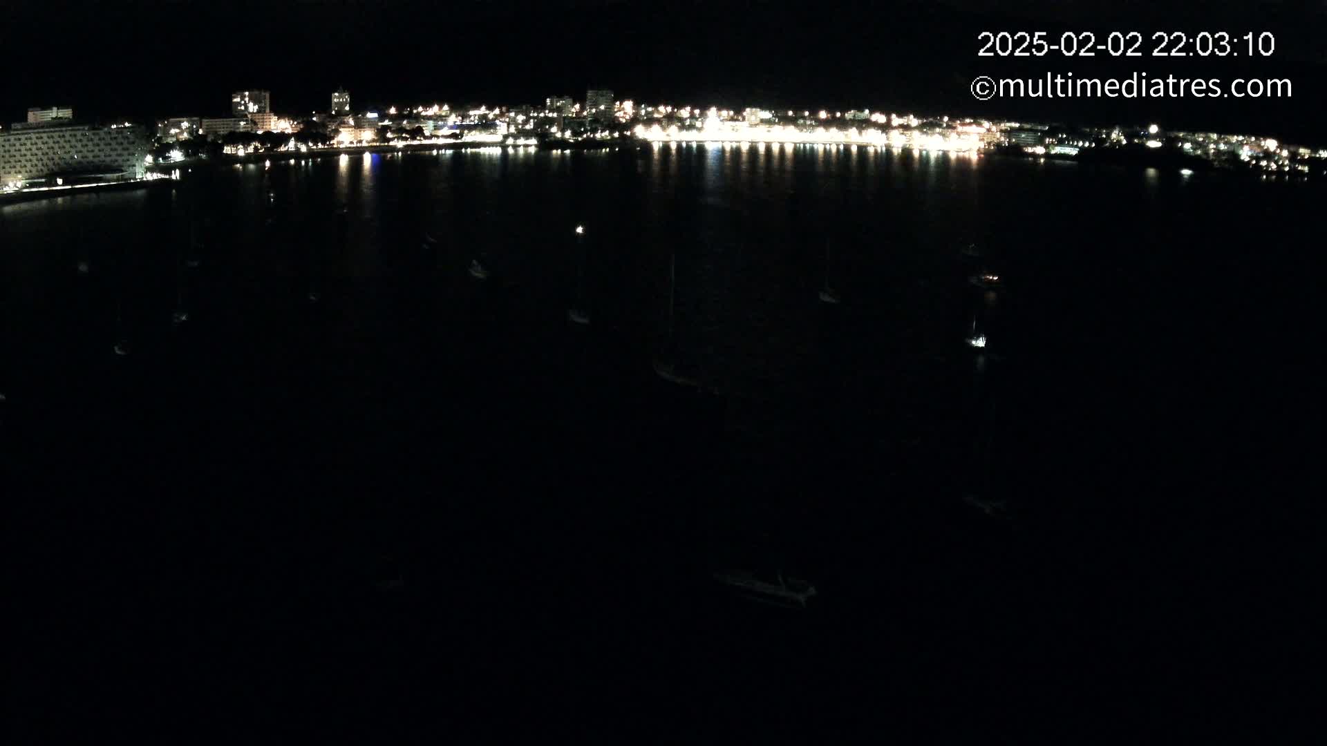 A nighttime, dark view of a city's waterfront at night, with many small lights on boats in dark water, and a brightly lit city skyline in the distance.