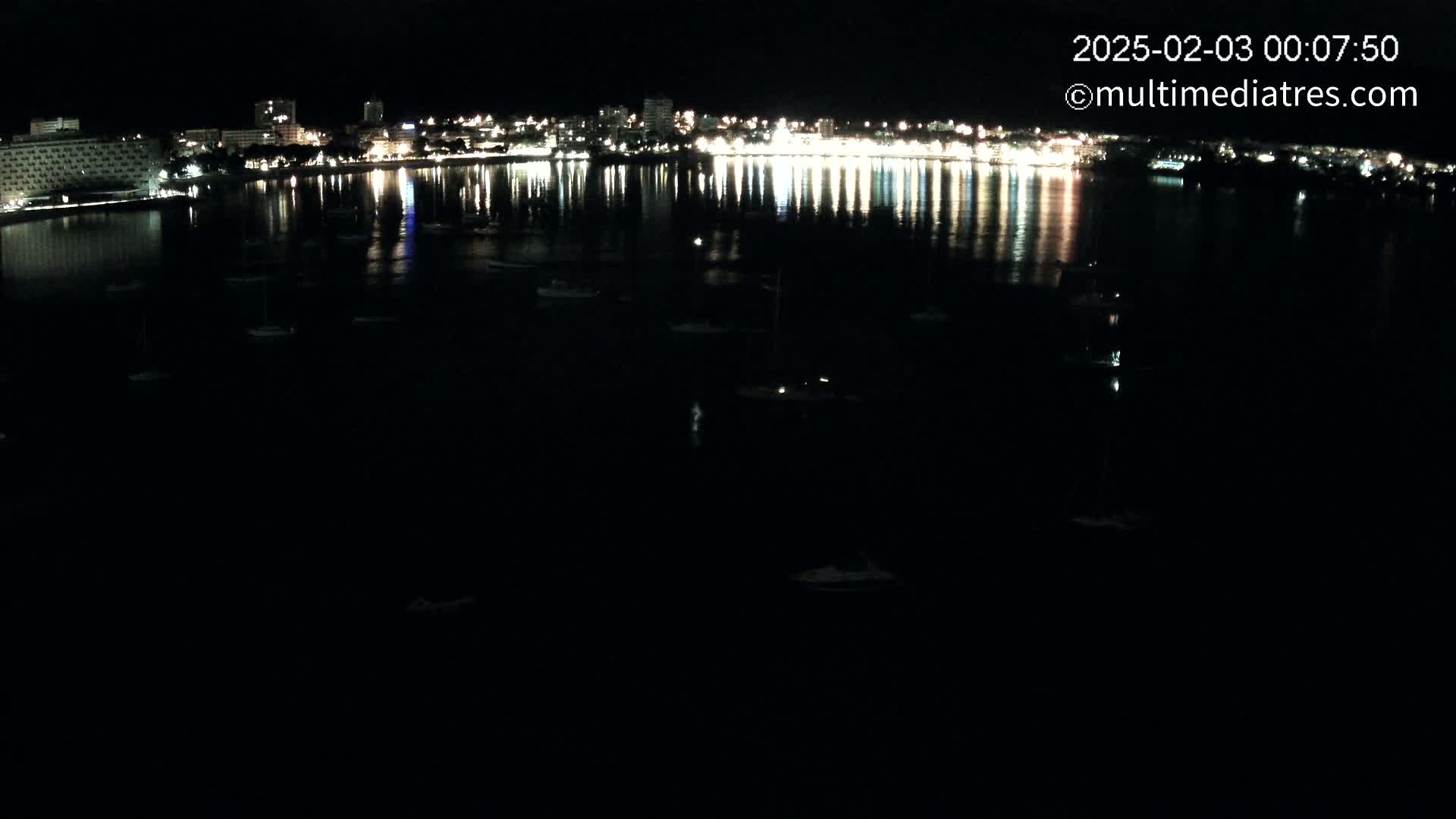 A nighttime view of a city's illuminated waterfront, reflecting in a dark body of water dotted with small boats.