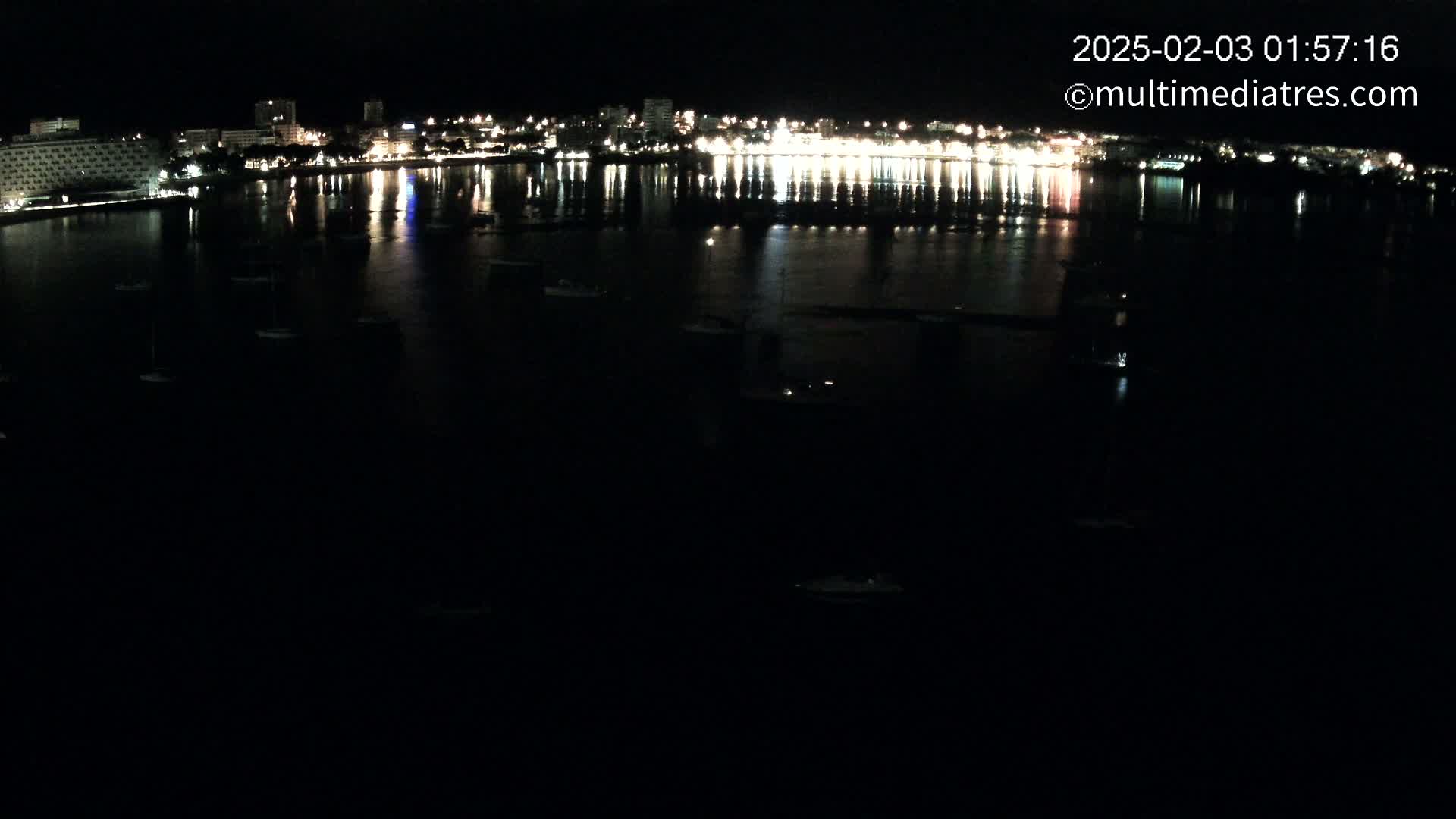 At night, a calm body of water reflects the lights of a city skyline, dotted with small boats.