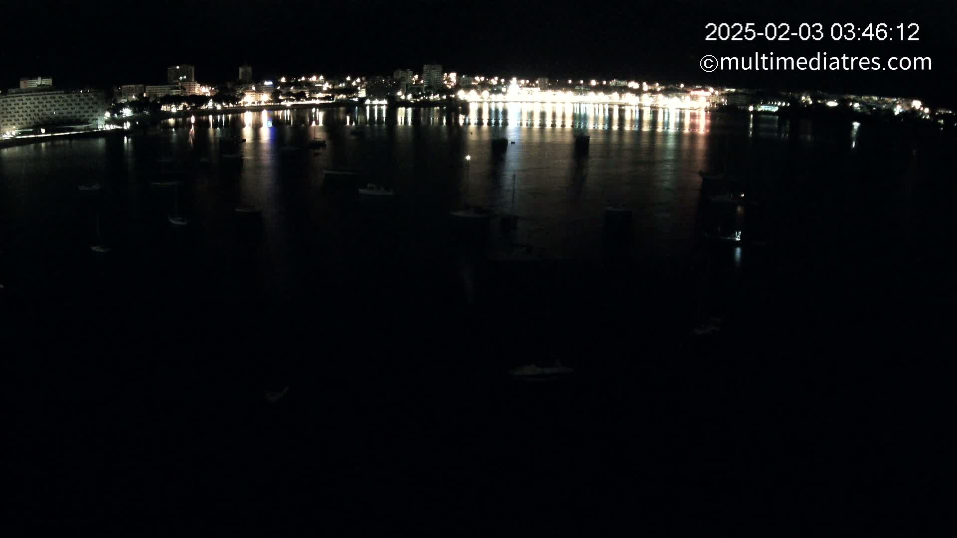 A nighttime view of a calm body of water with numerous small boats and a brightly lit city skyline reflected on the water's surface.