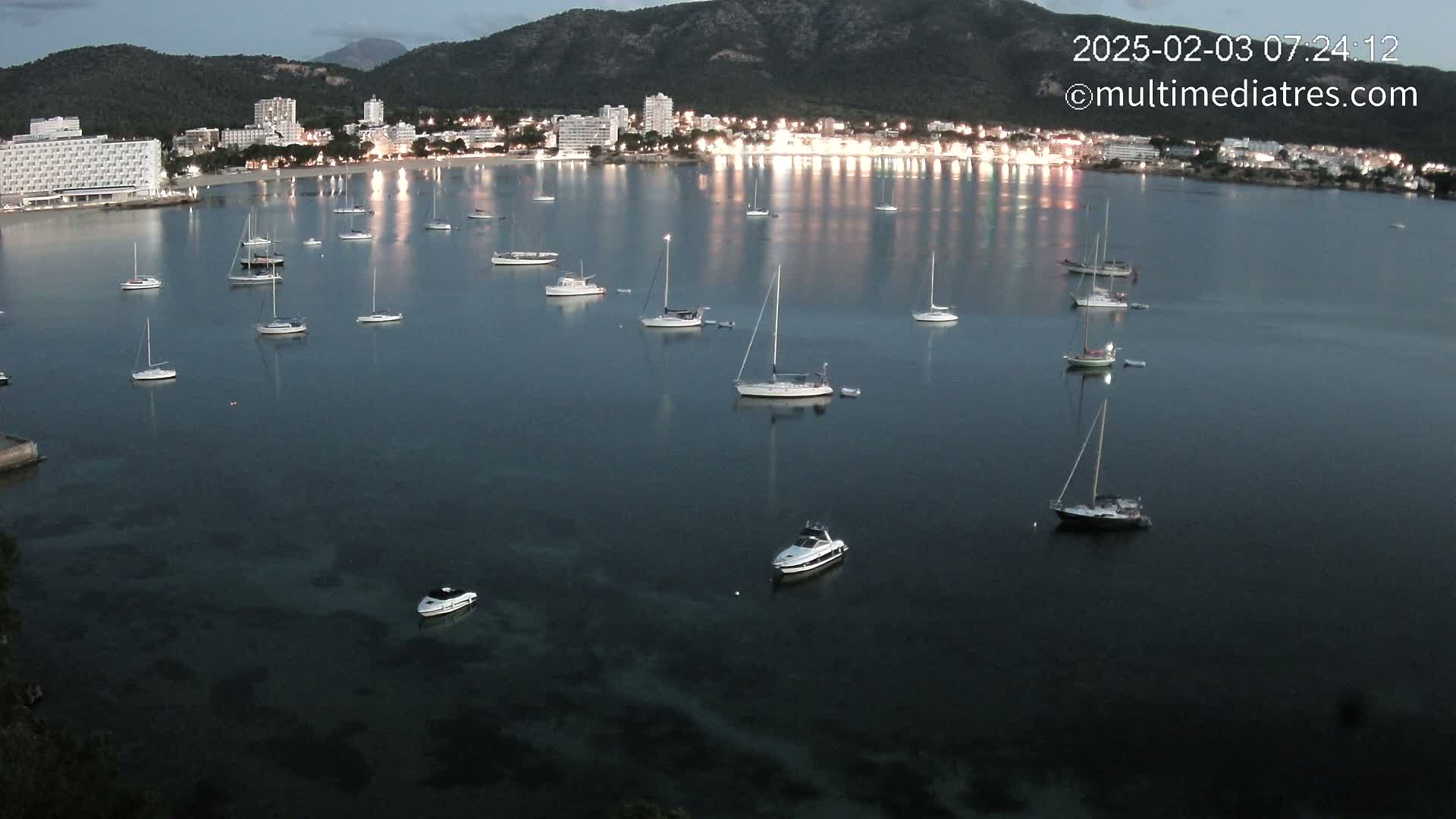 Numerous sailboats and motorboats are moored in a calm, dark-blue bay at dusk, with a city of hotels and buildings lit up along the shore, backed by dark mountains.