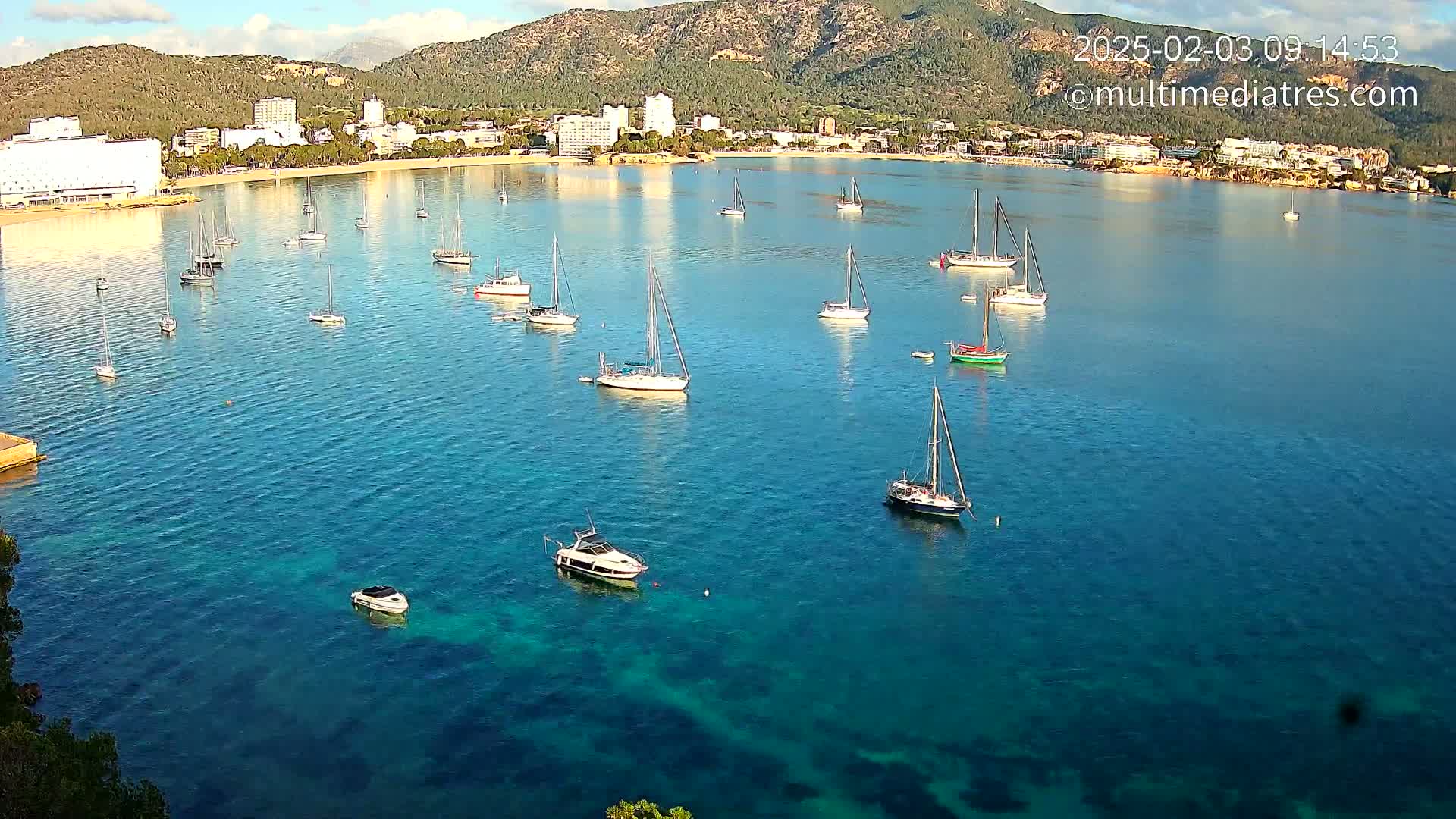 Numerous sailboats and motorboats are anchored in a calm, clear blue bay, with a town and mountains visible in the background under a mostly sunny sky.