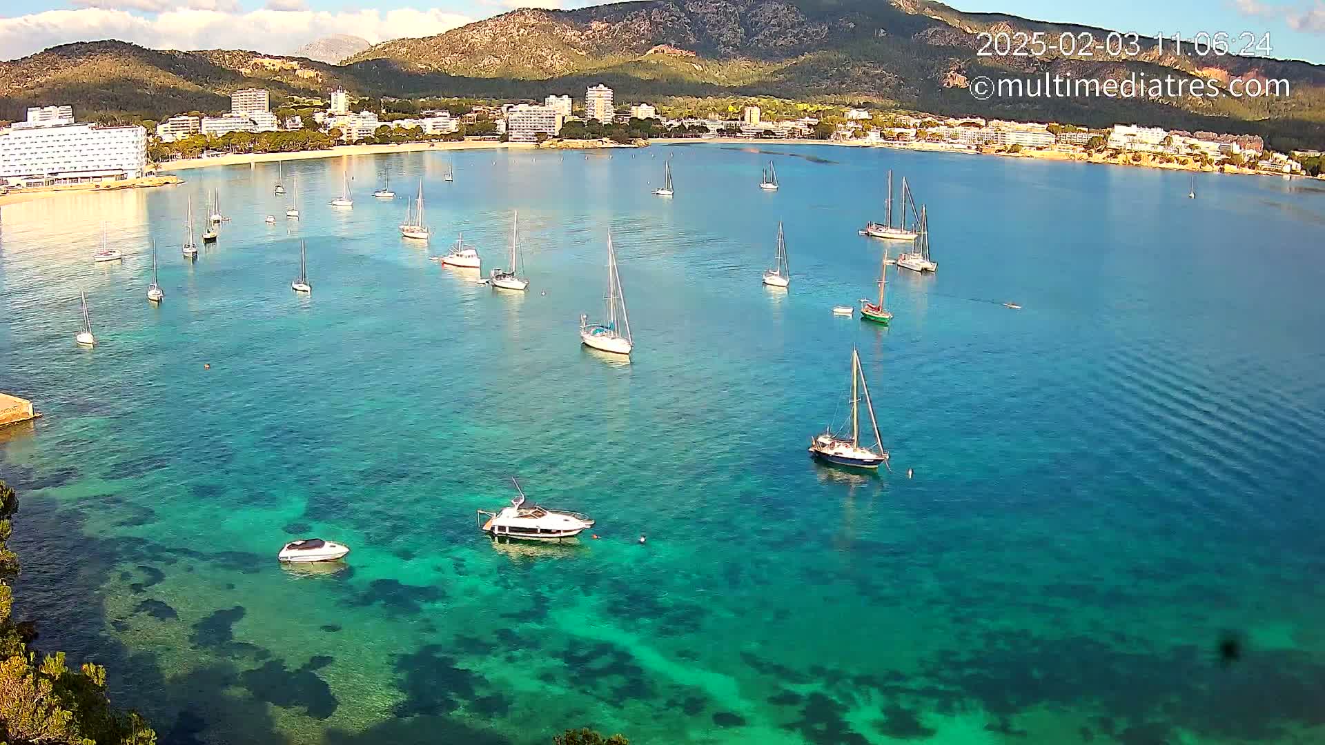 A calm, clear turquoise bay with numerous sailboats and motorboats anchored near a shoreline with hotels and mountains in the background.