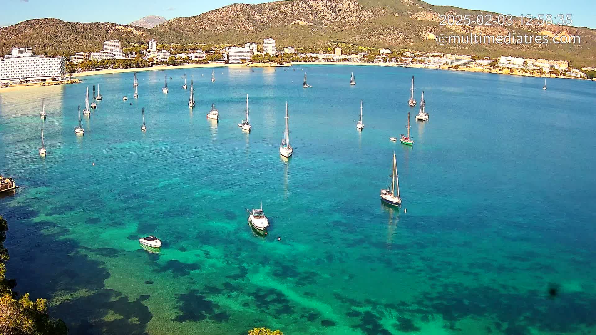 Numerous sailboats are anchored in a calm, clear turquoise bay, near a shoreline with buildings and a mountain in the background under sunny skies.