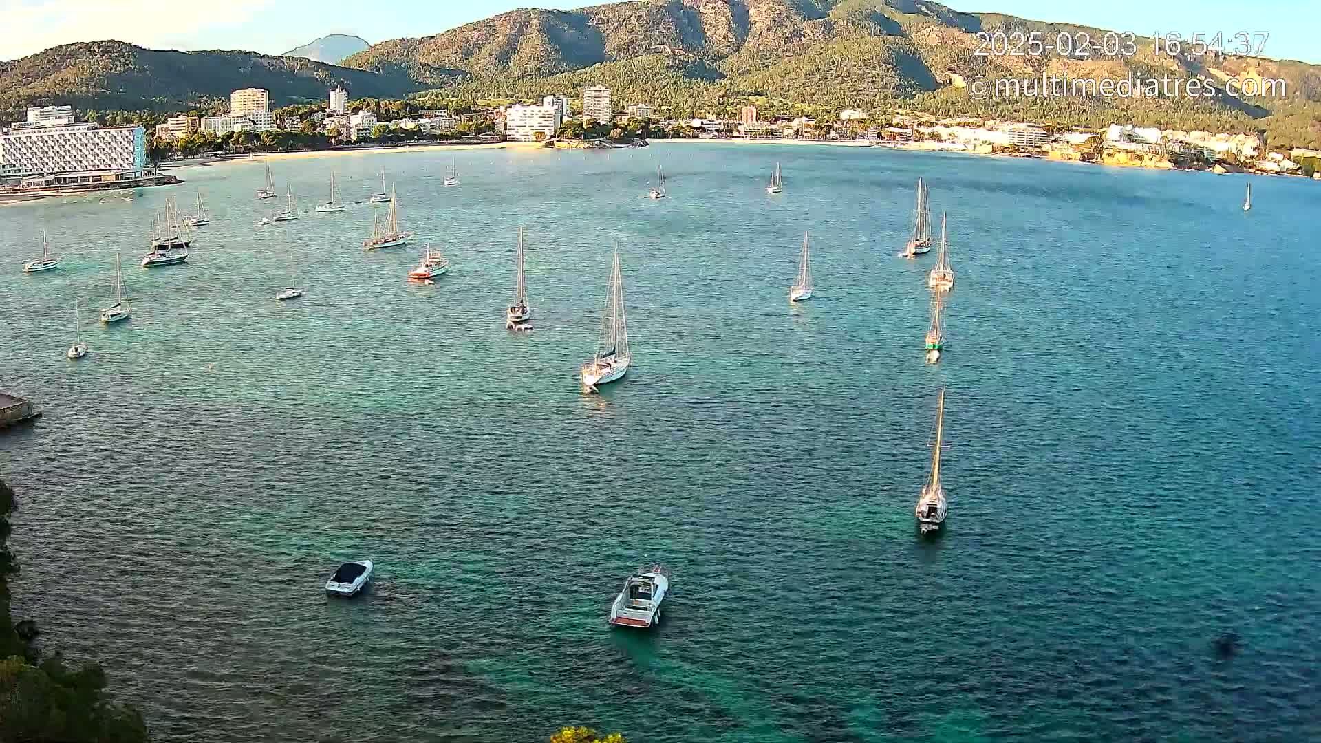 Numerous sailboats and motorboats are anchored in a calm, clear bay near a shoreline with buildings and mountains under a sunny sky.