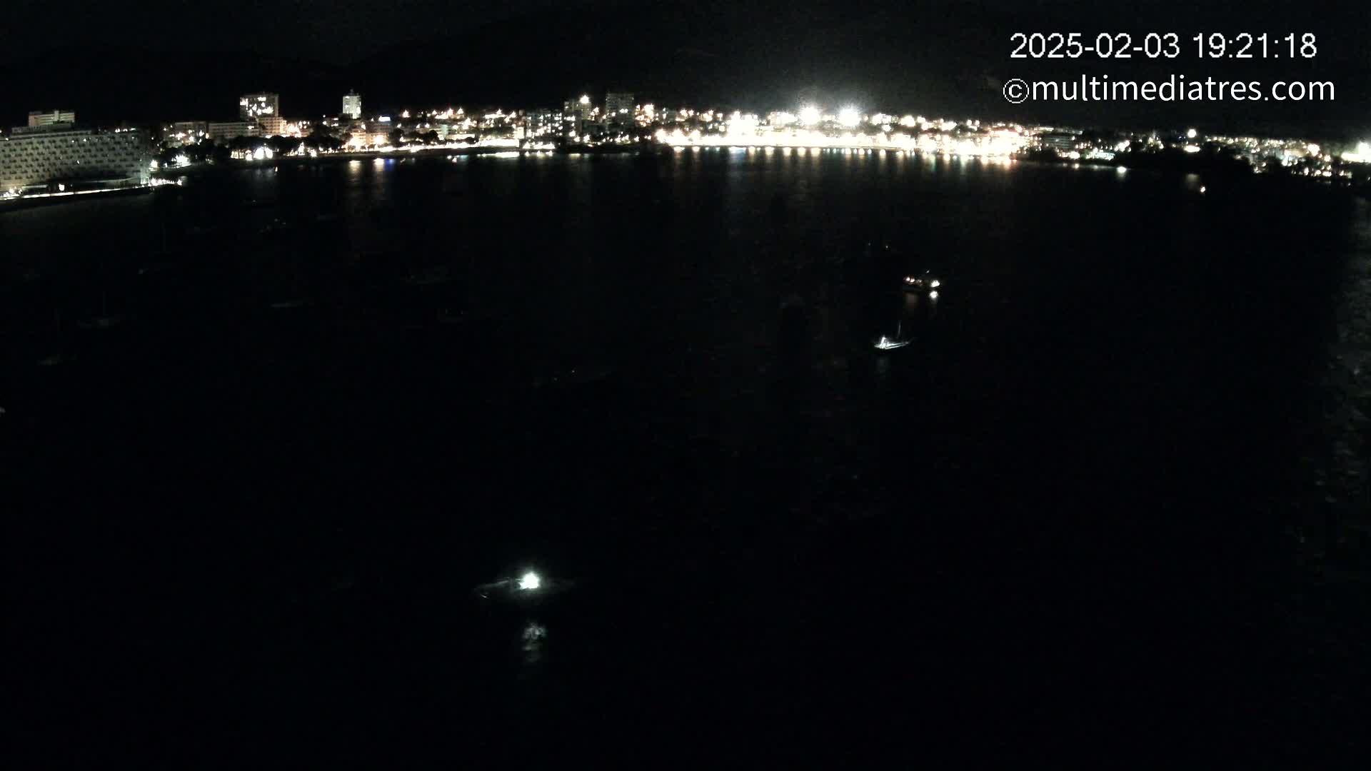 A nighttime, long shot of a dark body of water with a few boats, in front of a brightly lit city skyline.