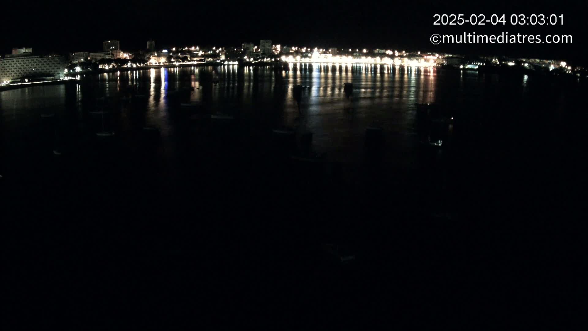 A nighttime view of a city's waterfront at night, showing calm water reflecting the lights of buildings along the shore.