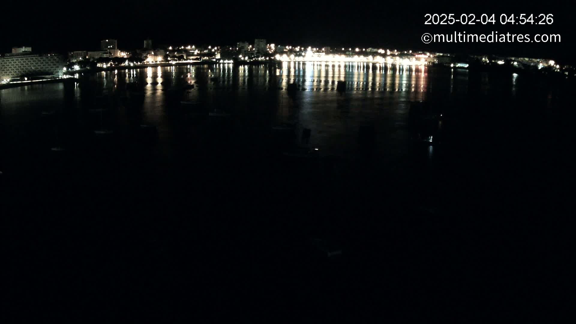A nighttime view of a calm body of water reflecting the lights of a city skyline across its surface, with several boats sparsely scattered.