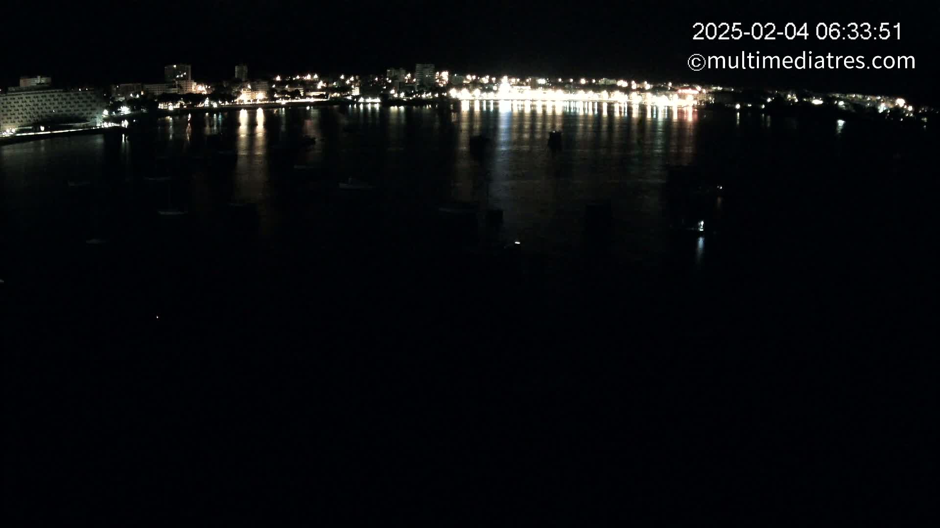 A nighttime view of a city's illuminated waterfront, with calm water reflecting the lights and several small boats scattered across the bay.