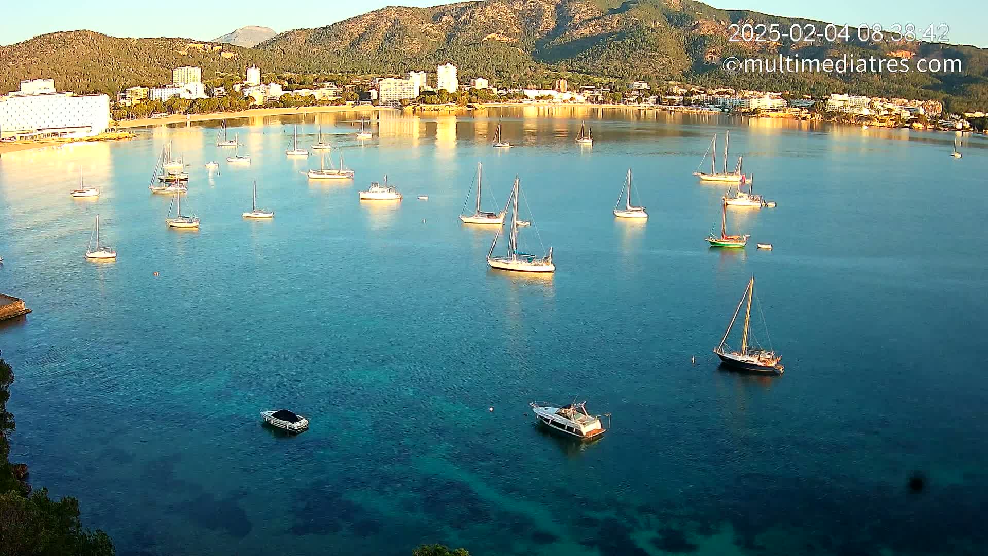 Numerous sailboats are anchored in a calm, clear bay near a shoreline with hotels and mountains under a bright, sunny sky.