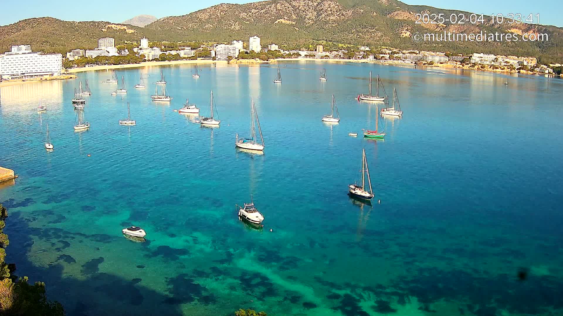 Numerous sailboats and motorboats are anchored in a calm, clear turquoise bay, with a mountainous shoreline lined with buildings in the background under a bright sunny sky.