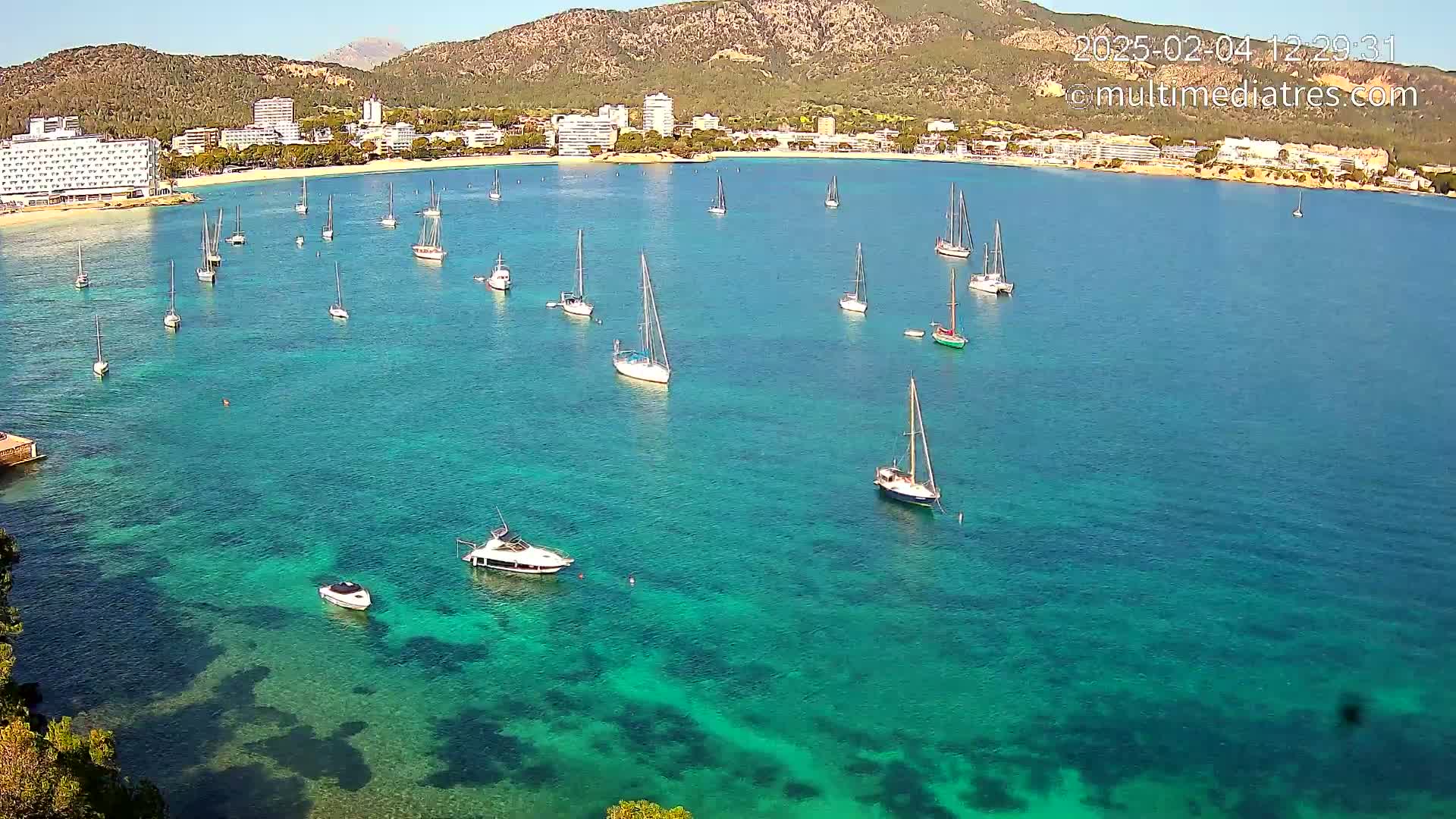 An aerial view shows numerous sailboats and motorboats in a calm, clear turquoise bay, with a beach and hotels visible on the shore, backed by mountains under a sunny sky.