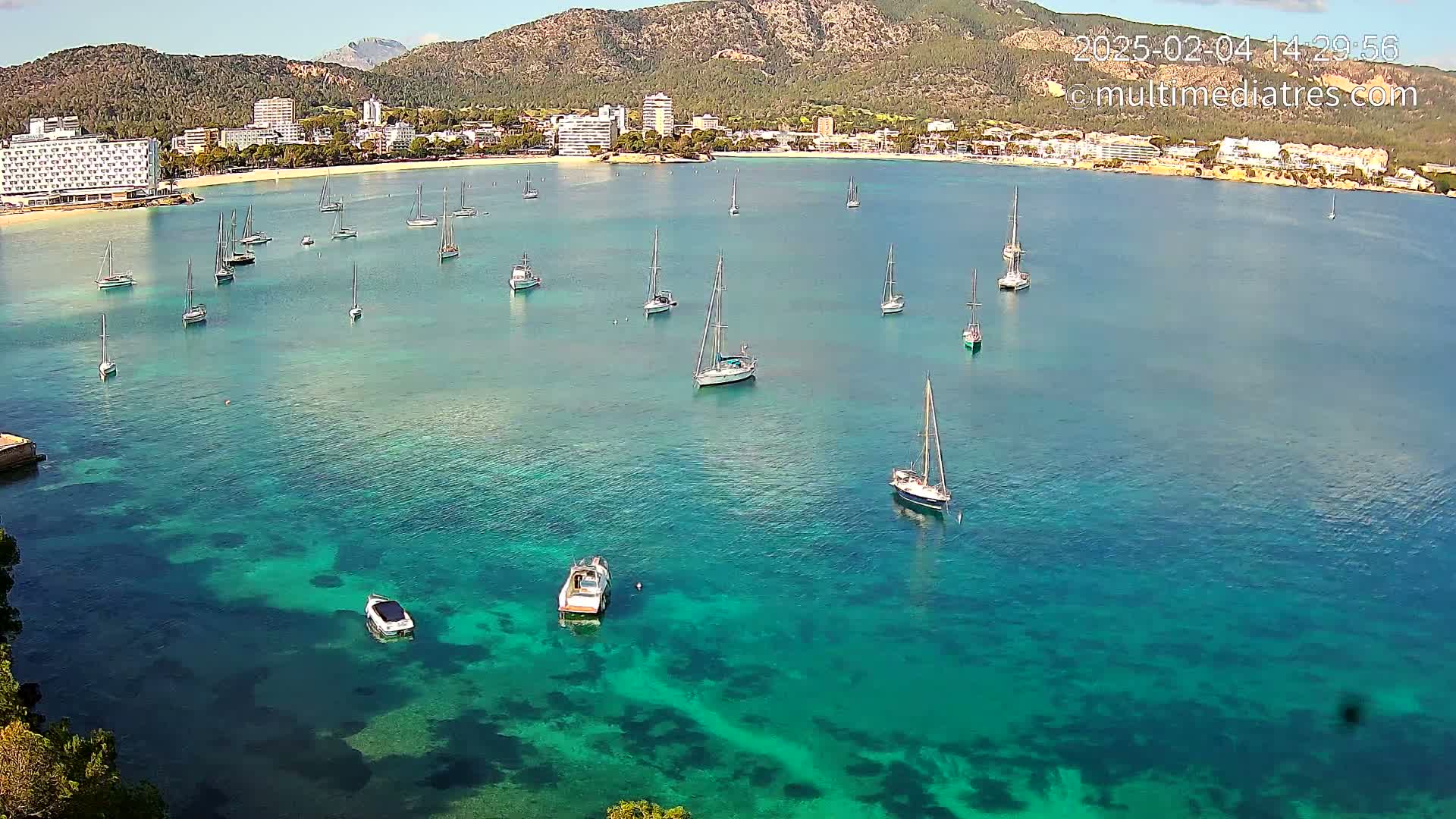A calm, sunny day shows a clear turquoise bay with numerous sailboats and motorboats anchored near a shoreline of hotels and mountains.