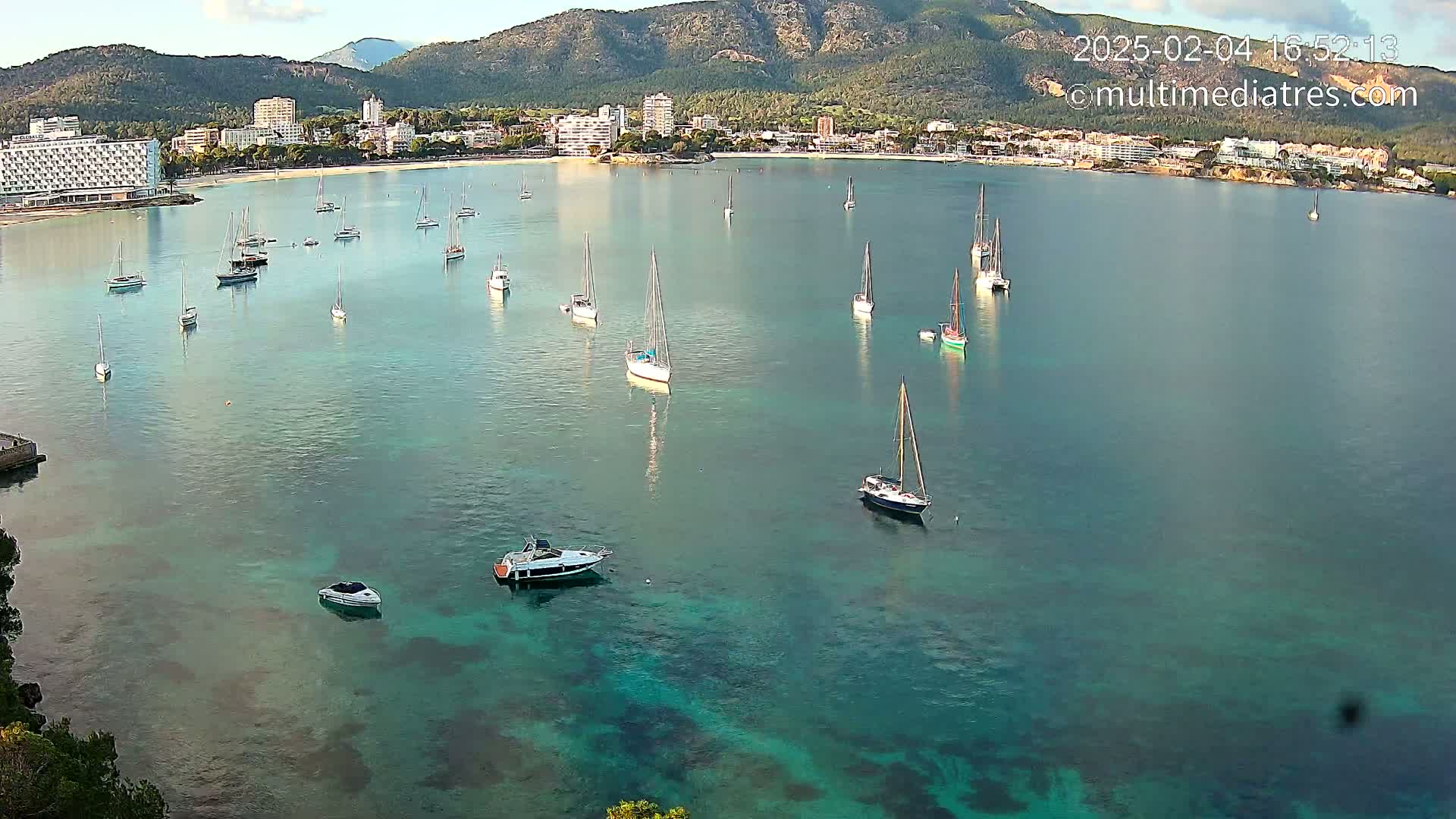 Numerous sailboats and motorboats are anchored in a calm, clear turquoise bay, with a coastal town and mountains visible in the background under a mostly sunny sky.