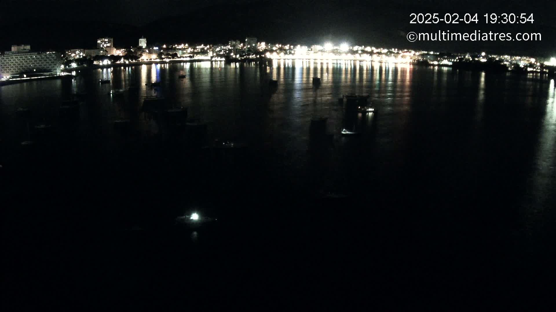A nighttime view of a calm body of water with numerous boats moored, reflecting the lights of a brightly lit city across the water.