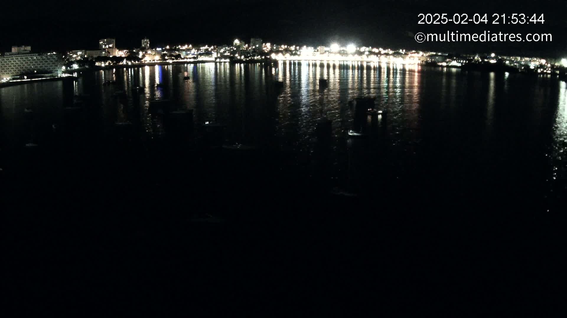 A nighttime view of a city's illuminated waterfront, reflecting in calm water, with several small boats visible in the bay.