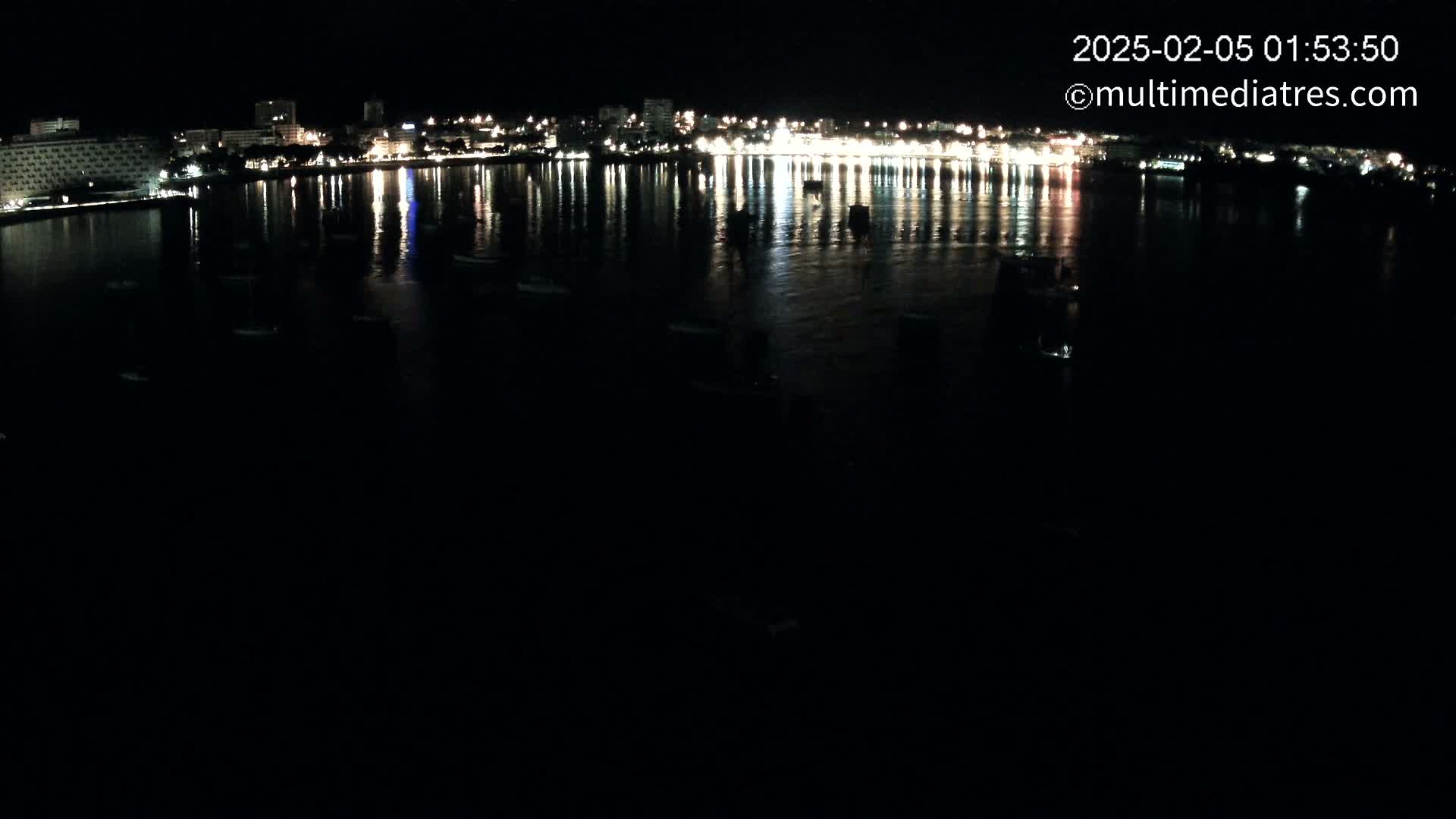 A nighttime view of a city's waterfront, showing its lights reflected in a calm body of water where several boats are moored.