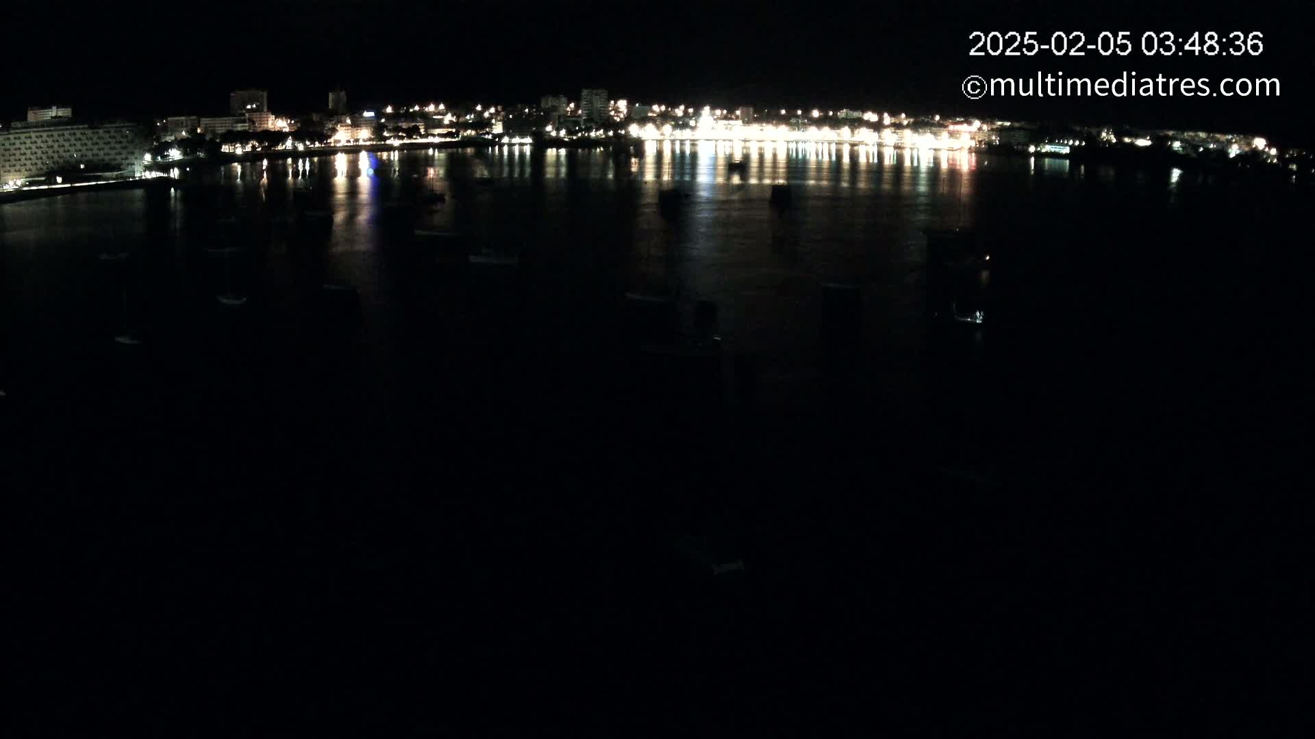 A nighttime view of a city's waterfront, showing a dark body of water with several small boats and a brightly lit city skyline in the distance.